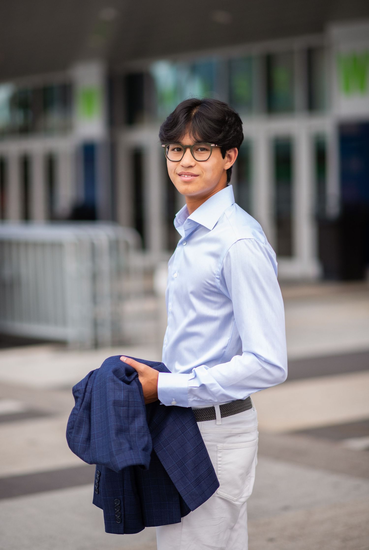 Young man in light blue dress shirt holding navy blazer outdoors in urban setting.