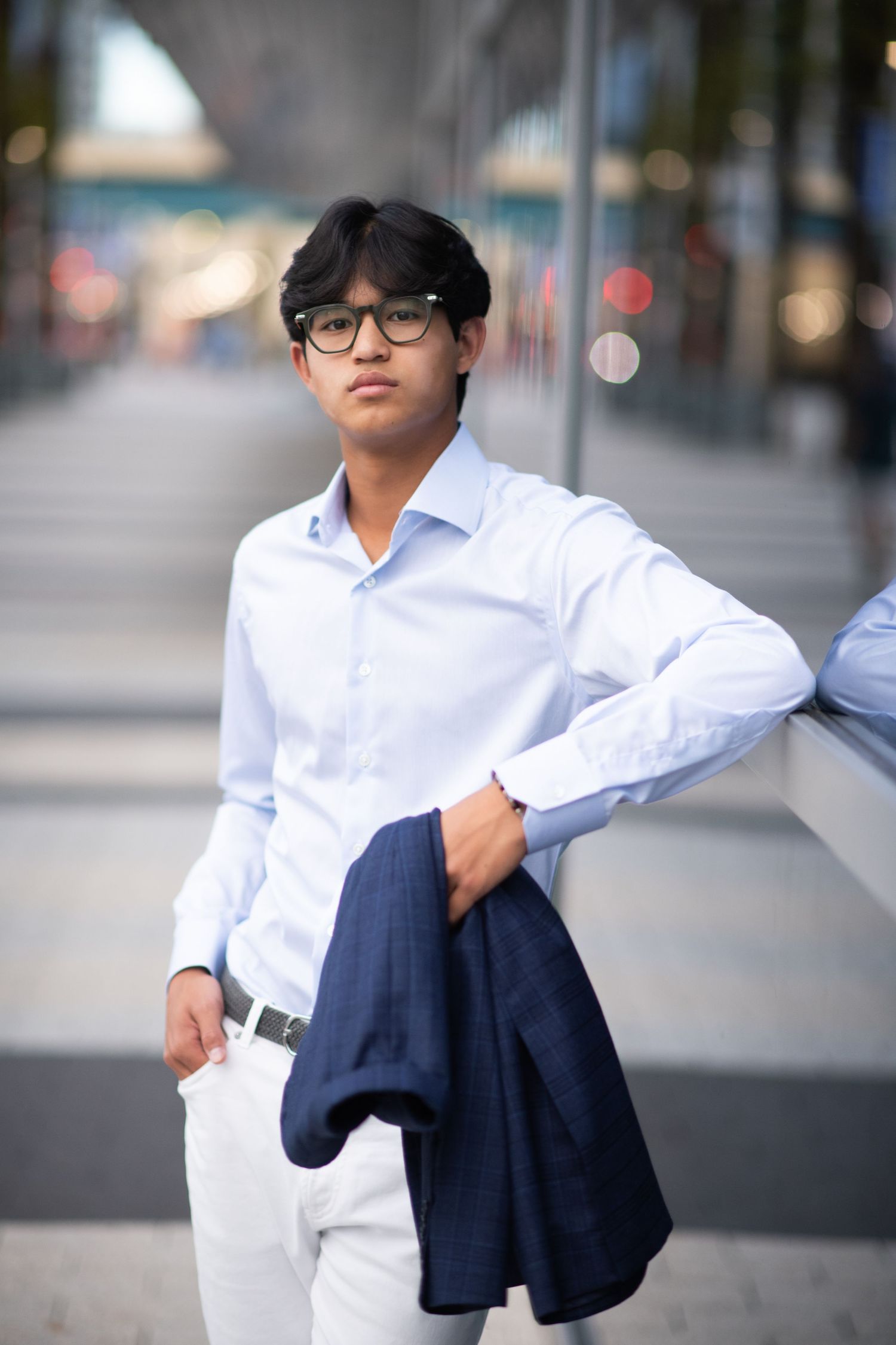Stylish young man in blue shirt and white pants leaning against wall holding blazer.