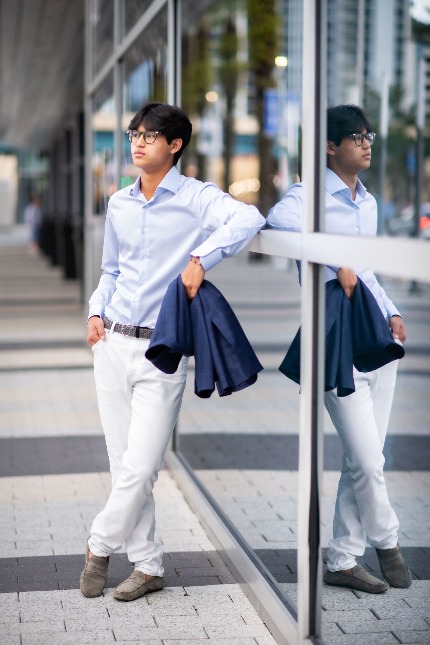 Stylish young Asian man in blue shirt and white pants leaning on glass wall with reflection.