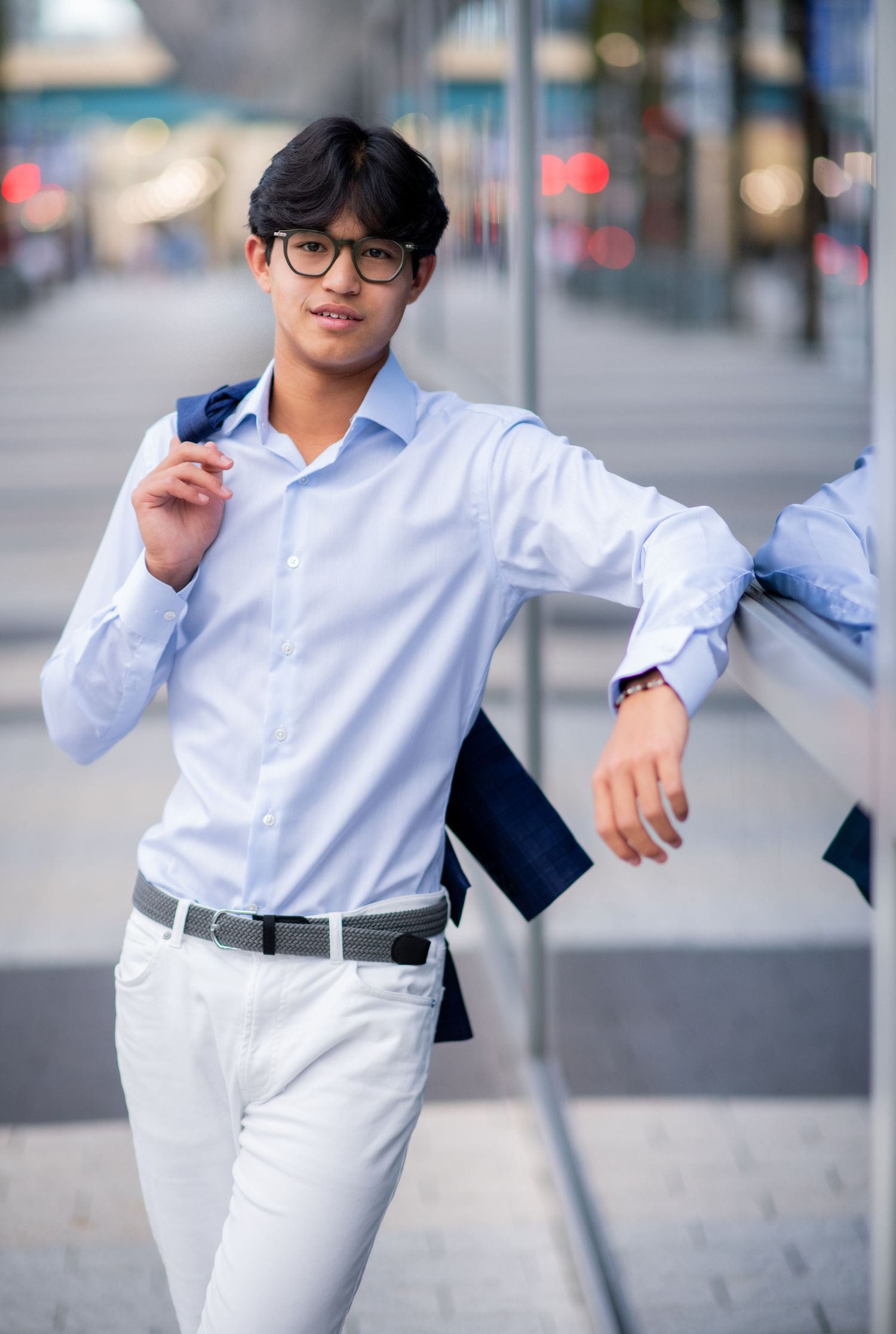 Young Asian man in blue blazer and white pants leaning on glass wall.