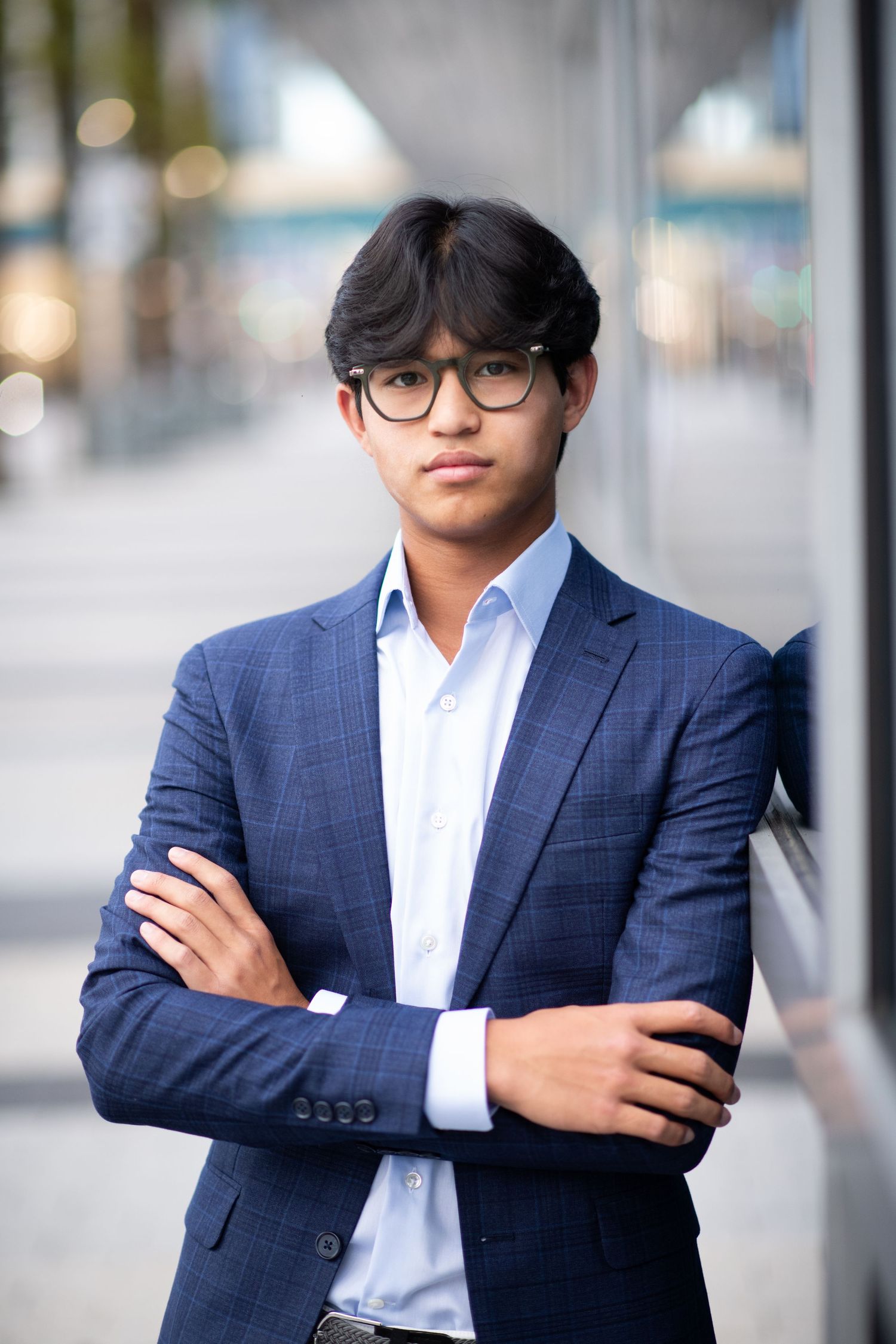Young Asian man in navy suit with arms crossed leaning on glass wall.