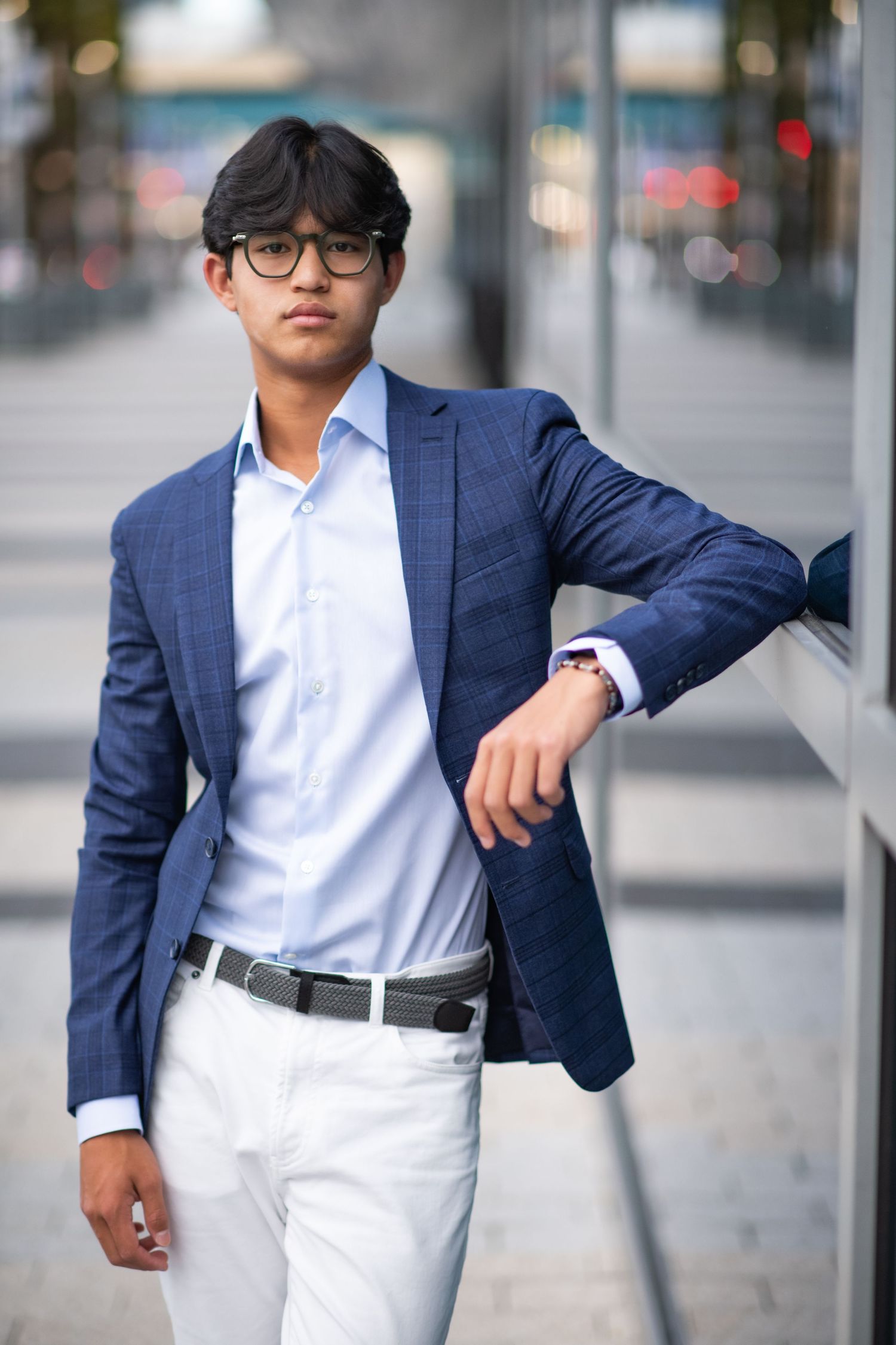 Confident young man in navy blazer and white pants leaning against glass wall outdoors.