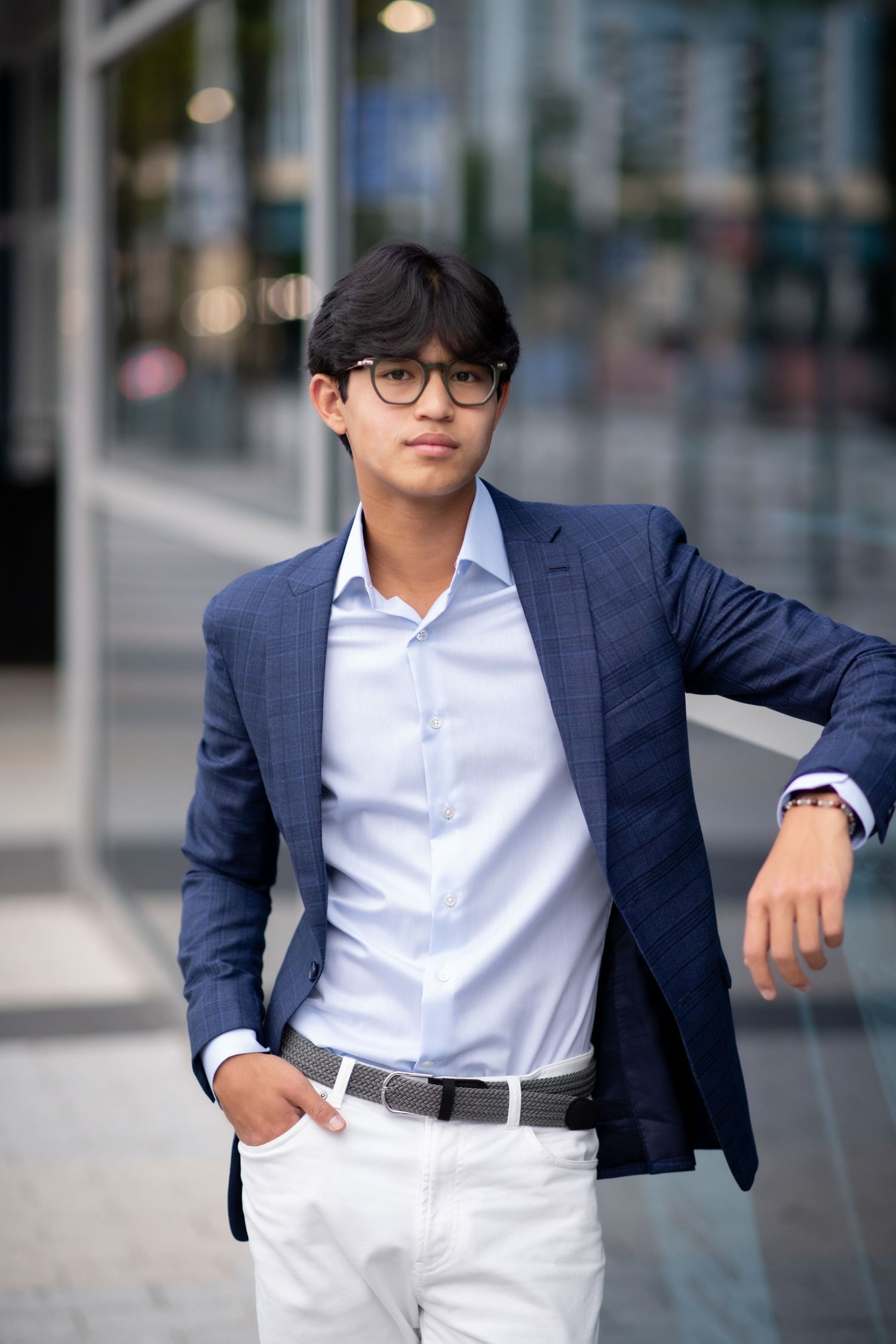 Young Asian man in navy blazer and white pants posing near glass building.