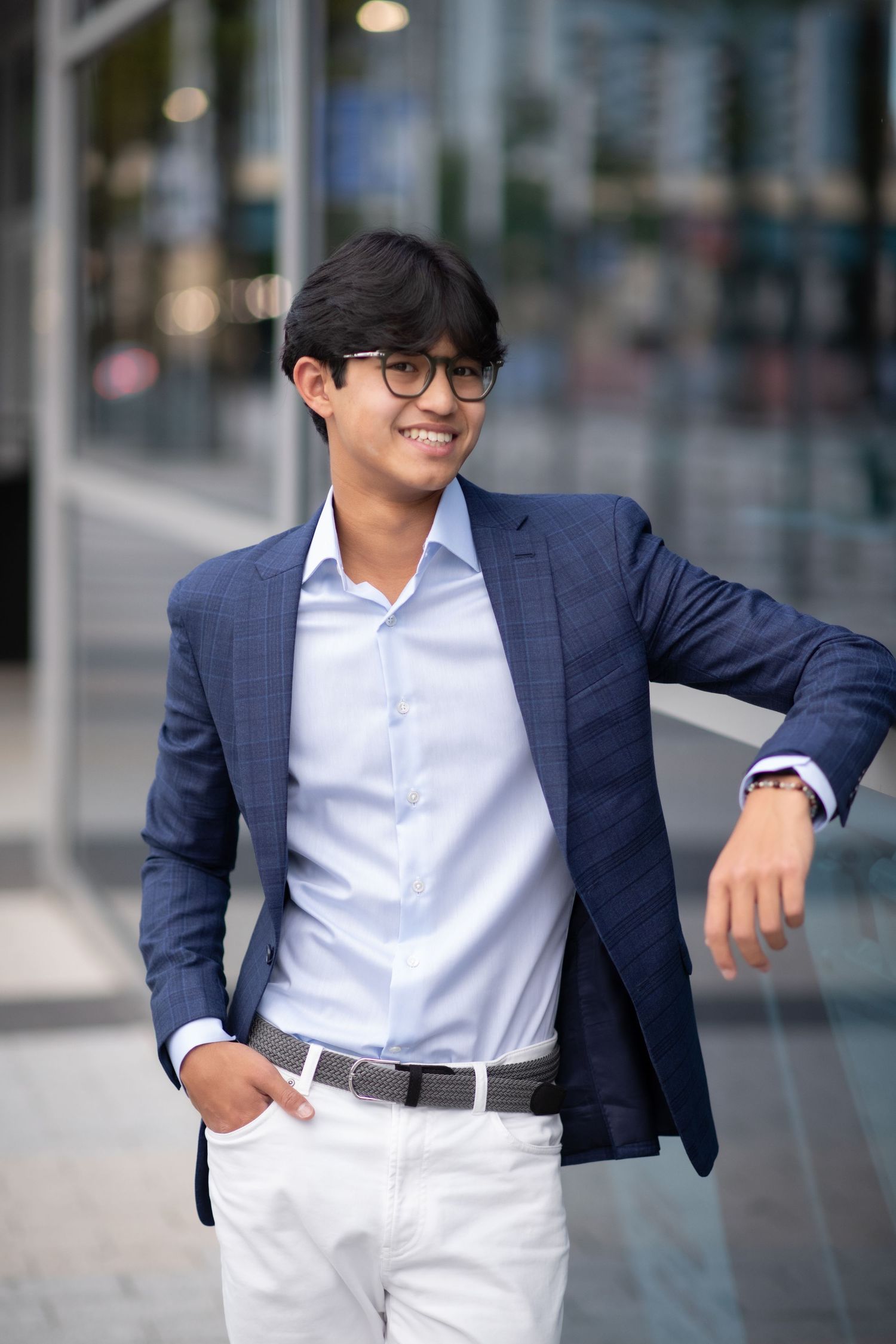 Young Asian businessman in navy blazer and glasses smiling outdoors near modern glass building.