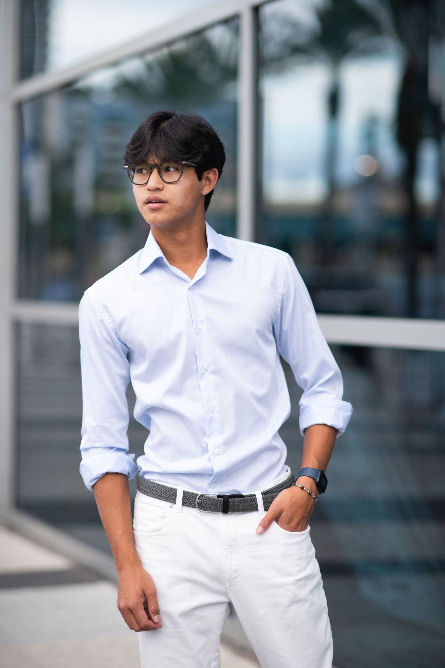 Young Asian man in light blue shirt, white pants, and glasses poses confidently outdoors near a modern glass building.