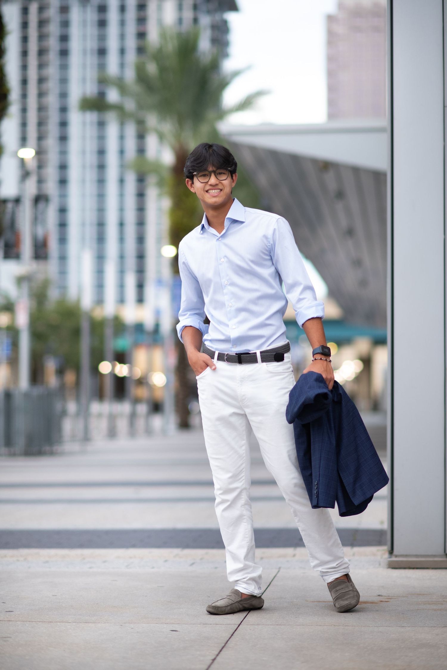 Smiling young Asian man in light blue shirt and white pants holding navy blazer outdoors in urban setting.