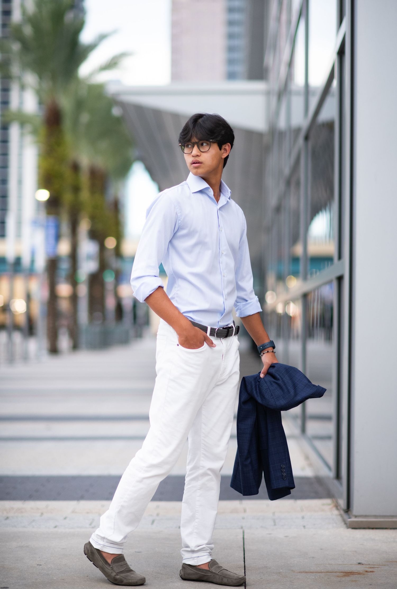 Stylish young Asian man walking in white pants and light blue shirt carrying a navy blazer in a city.