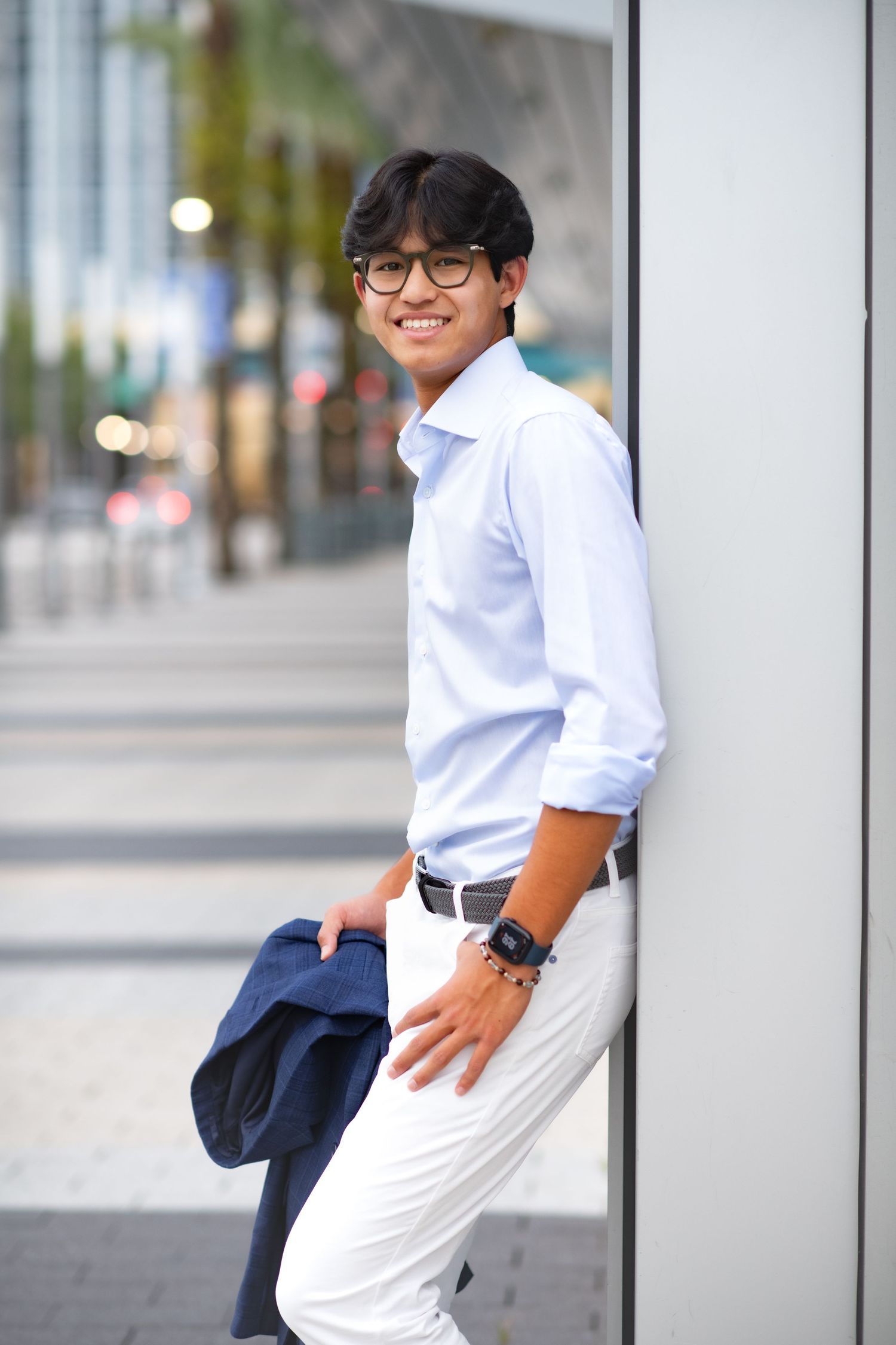 Smiling young Asian man in white shirt and pants, holding a jacket, leaning against a wall outdoors.