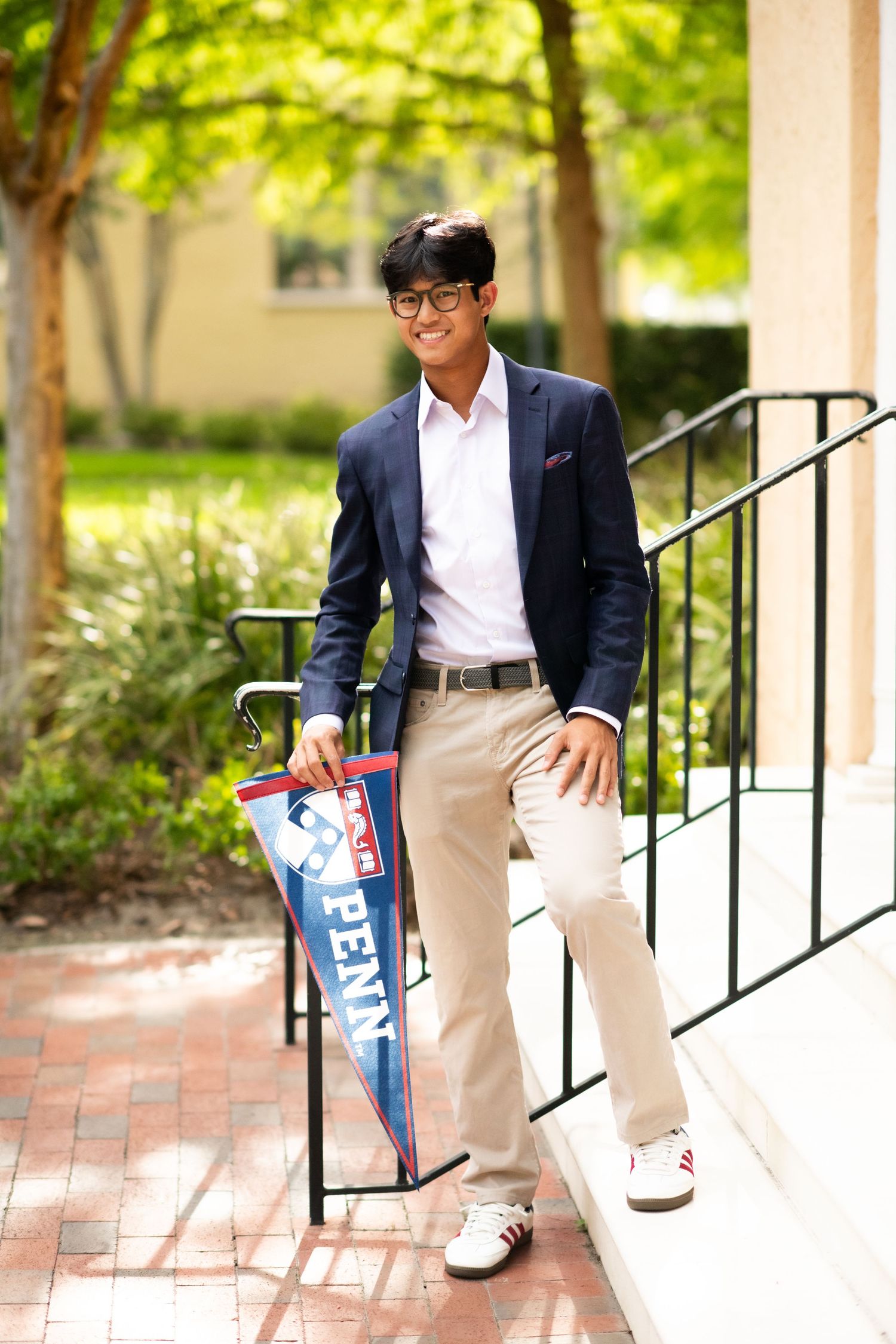 Young man in navy blazer holding a Penn pennant flag, standing on brick steps outdoors near green trees.