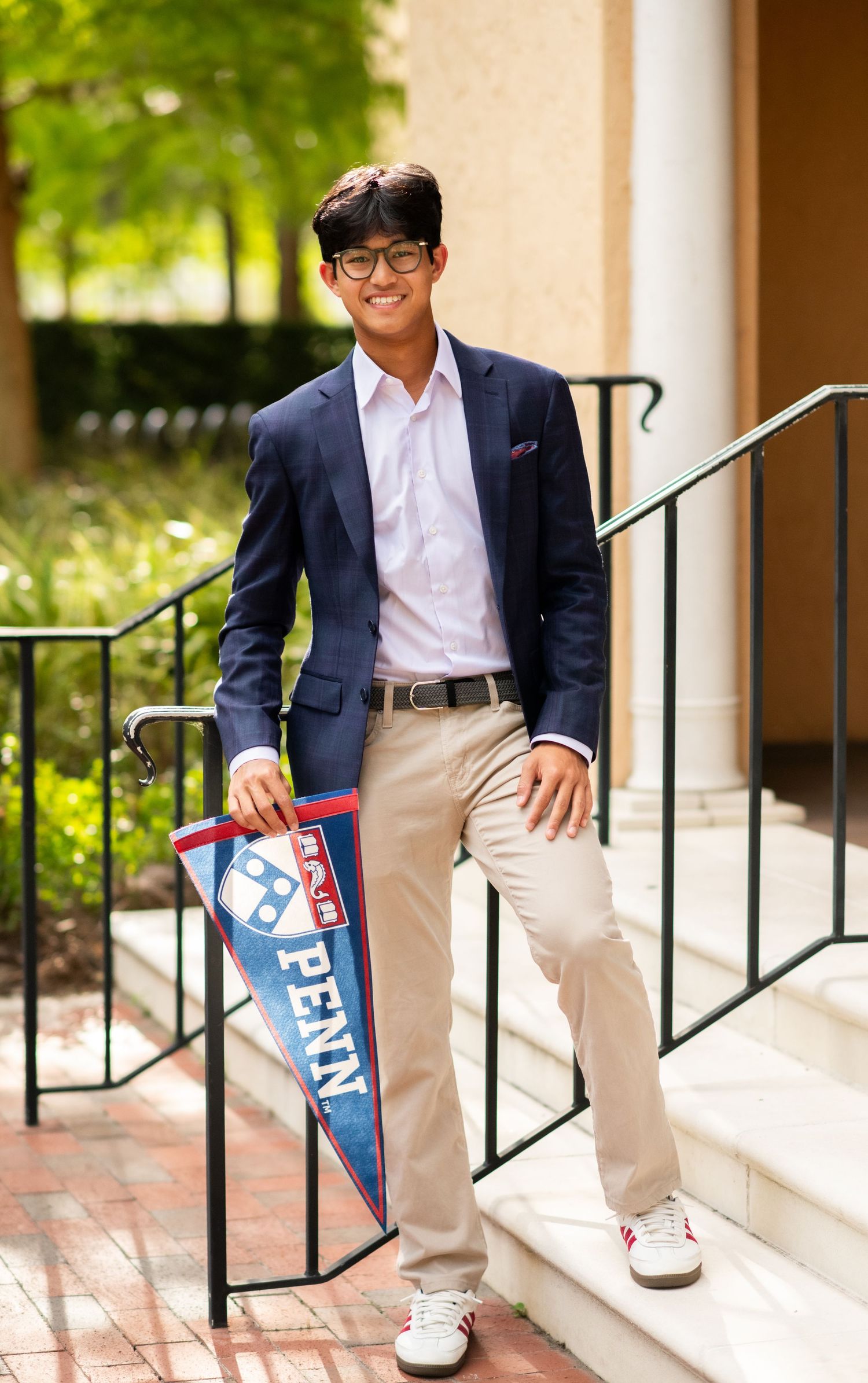 Young man in navy blazer and khakis holding a Penn pennant flag while smiling on outdoor steps.