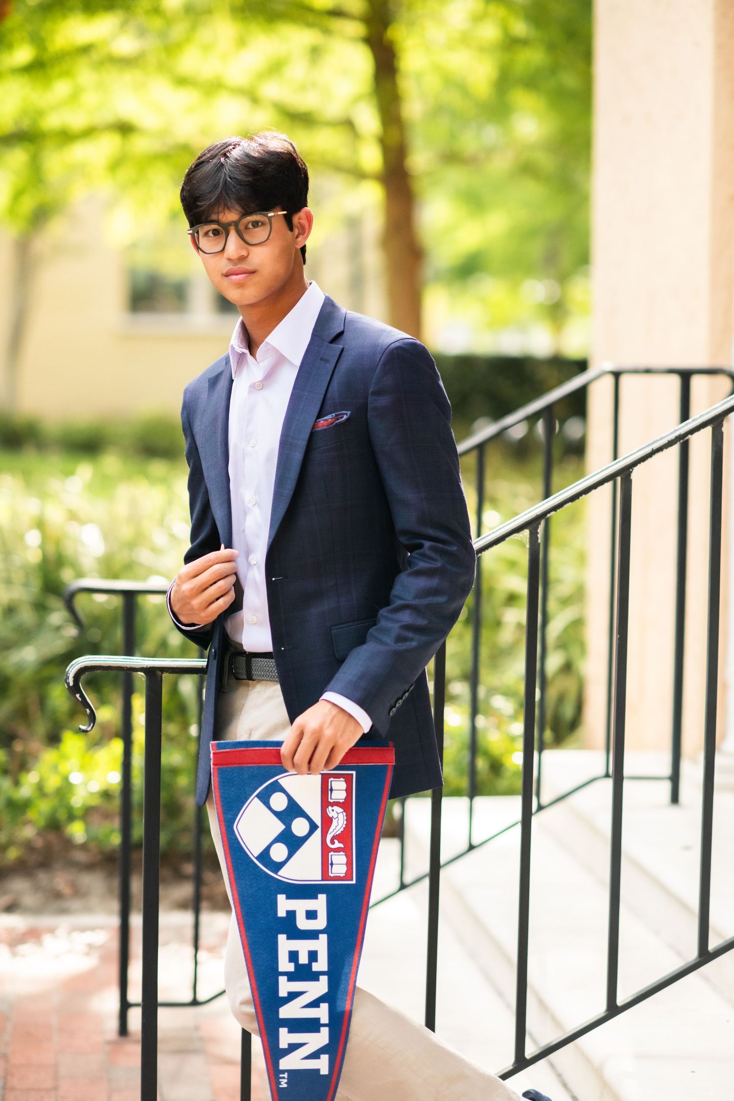 Young Asian man in navy blazer holding a Penn pennant on outdoor steps.