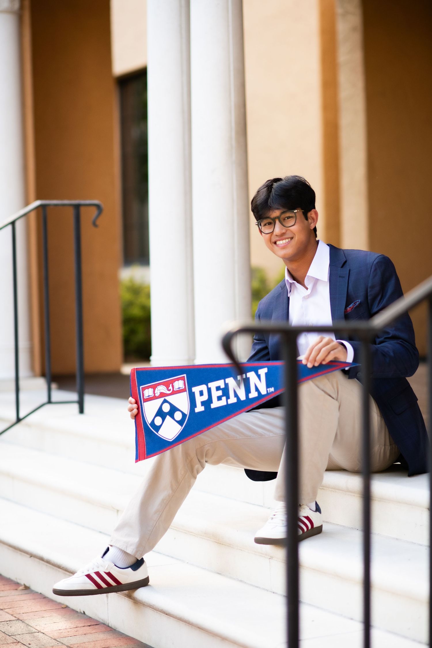 Smiling young man in navy blazer holding a University of Pennsylvania pennant while sitting on white steps.