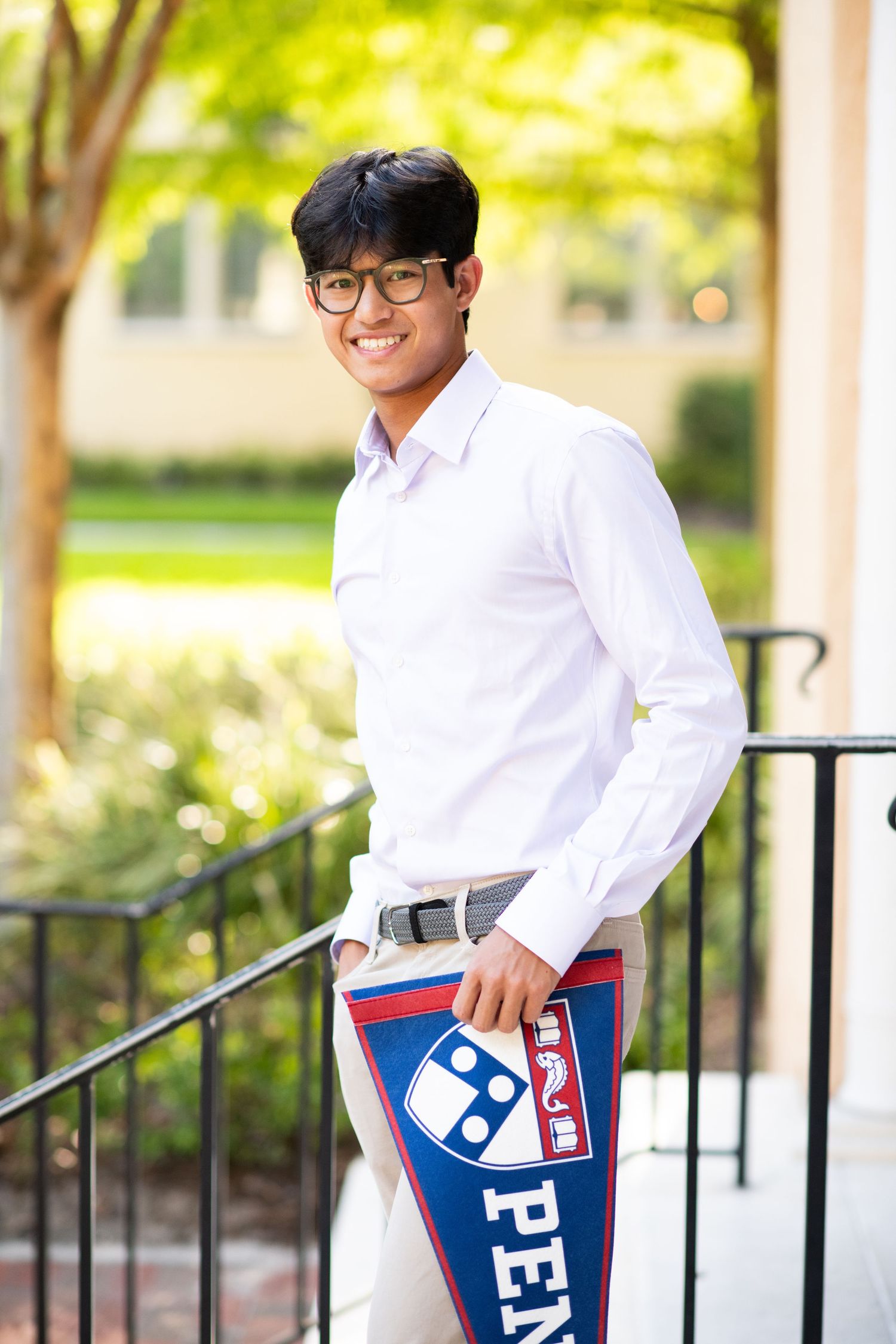 Smiling young man in white shirt holding a University of Pennsylvania pennant, standing by railing outdoors.