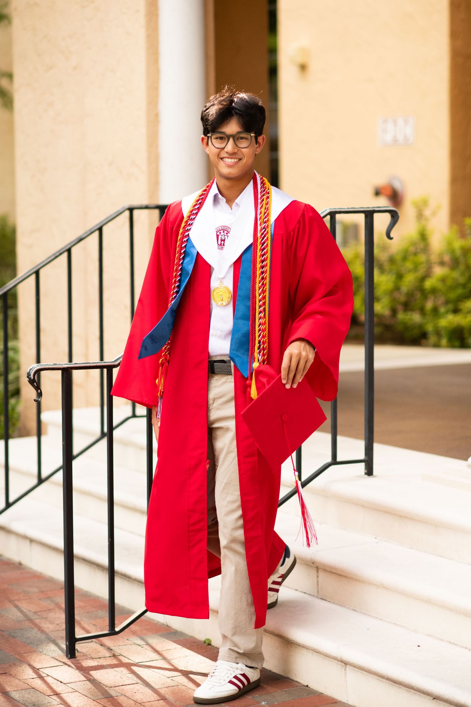 Young man in red graduation gown and cap standing on steps outdoors.