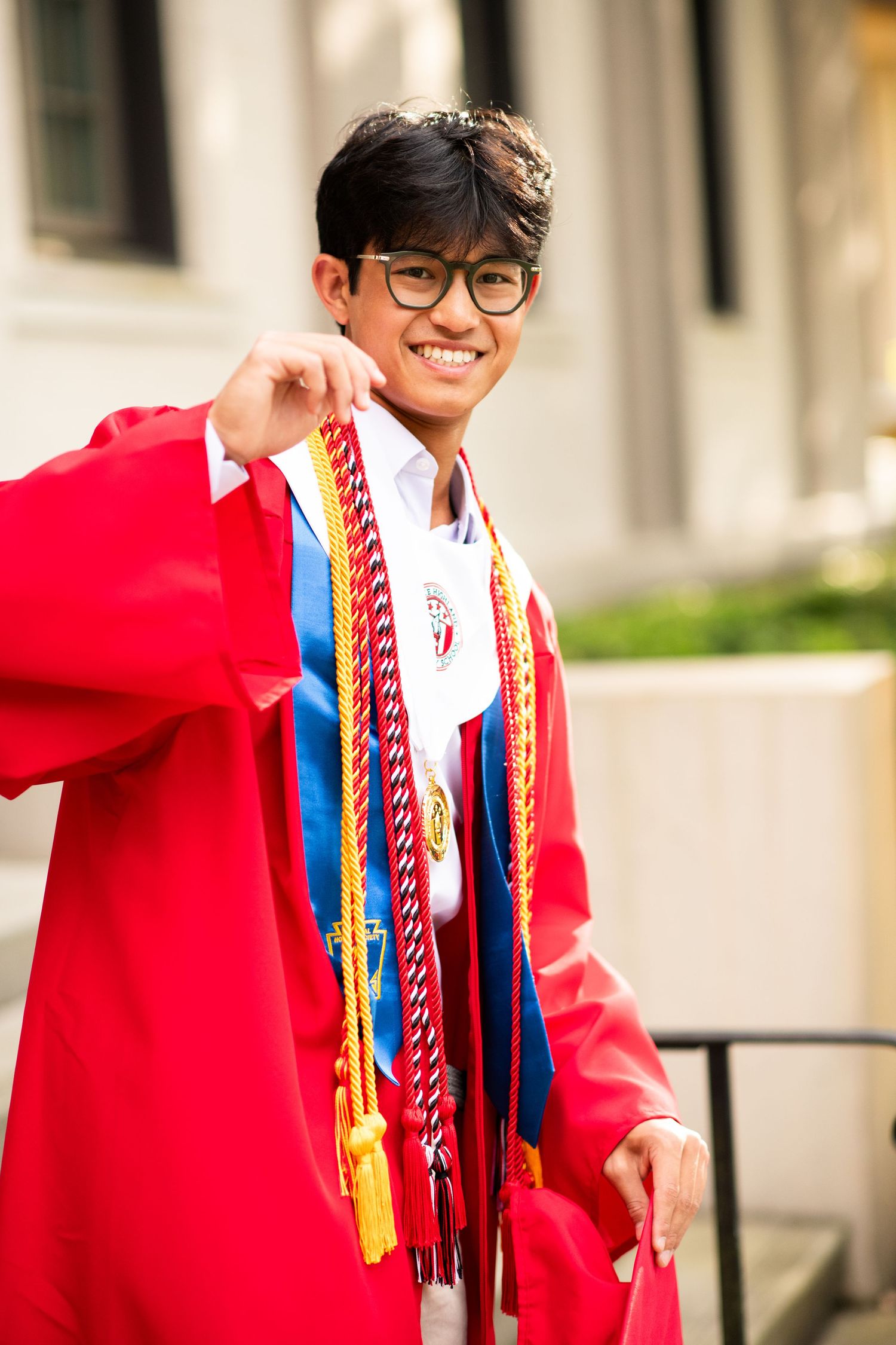 Happy graduate in red cap and gown with honor cords and sashes posing on outdoor steps.
