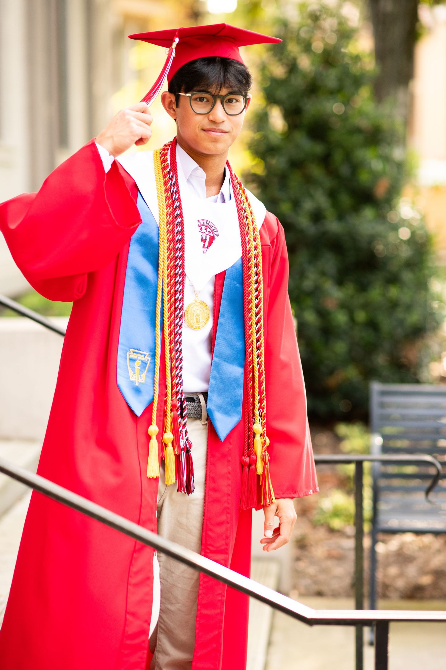 Young male graduate in red cap and gown with honor cords poses on outdoor steps.