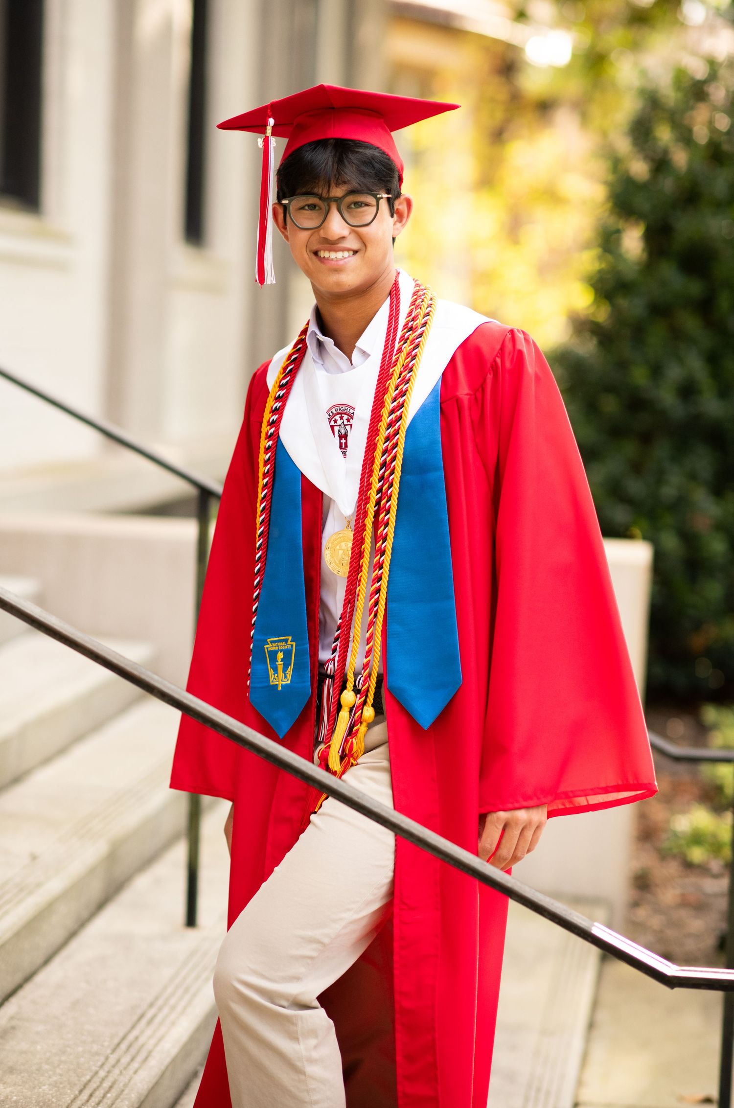 Smiling male graduate in red cap and gown with blue stole walks down outdoor steps.