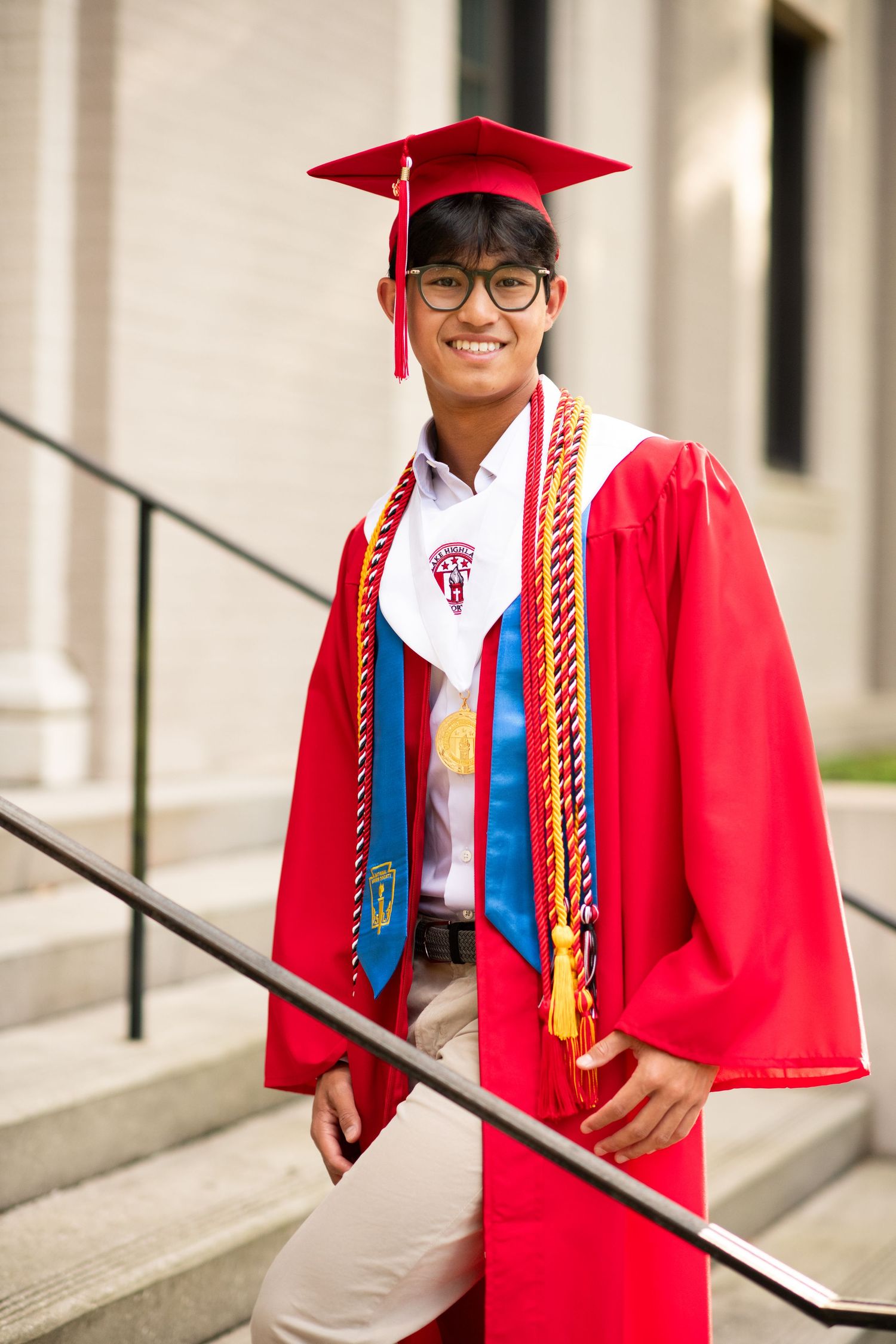 Smiling young man in red graduation cap and gown with honor cords standing on steps.