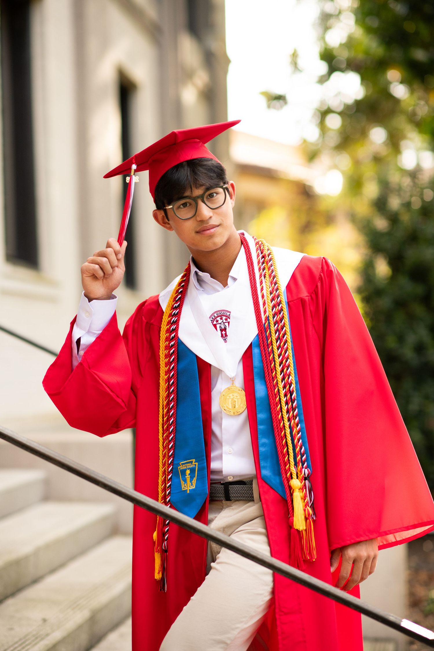 Male graduate in red cap and gown holding tassel poses on stone steps outdoors.
