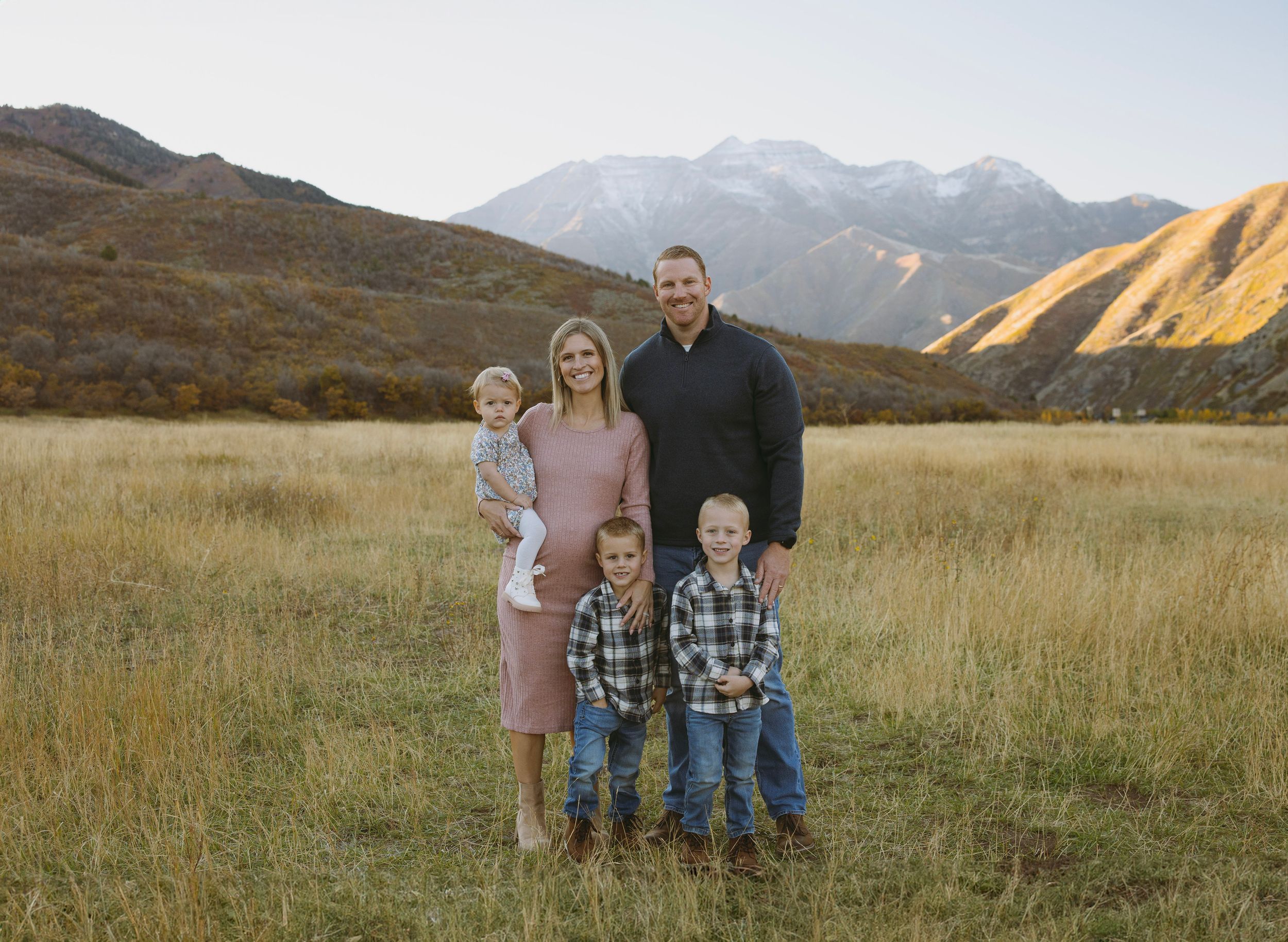 Family of five posing in a golden meadow with snow-capped mountains in the background during autumn.