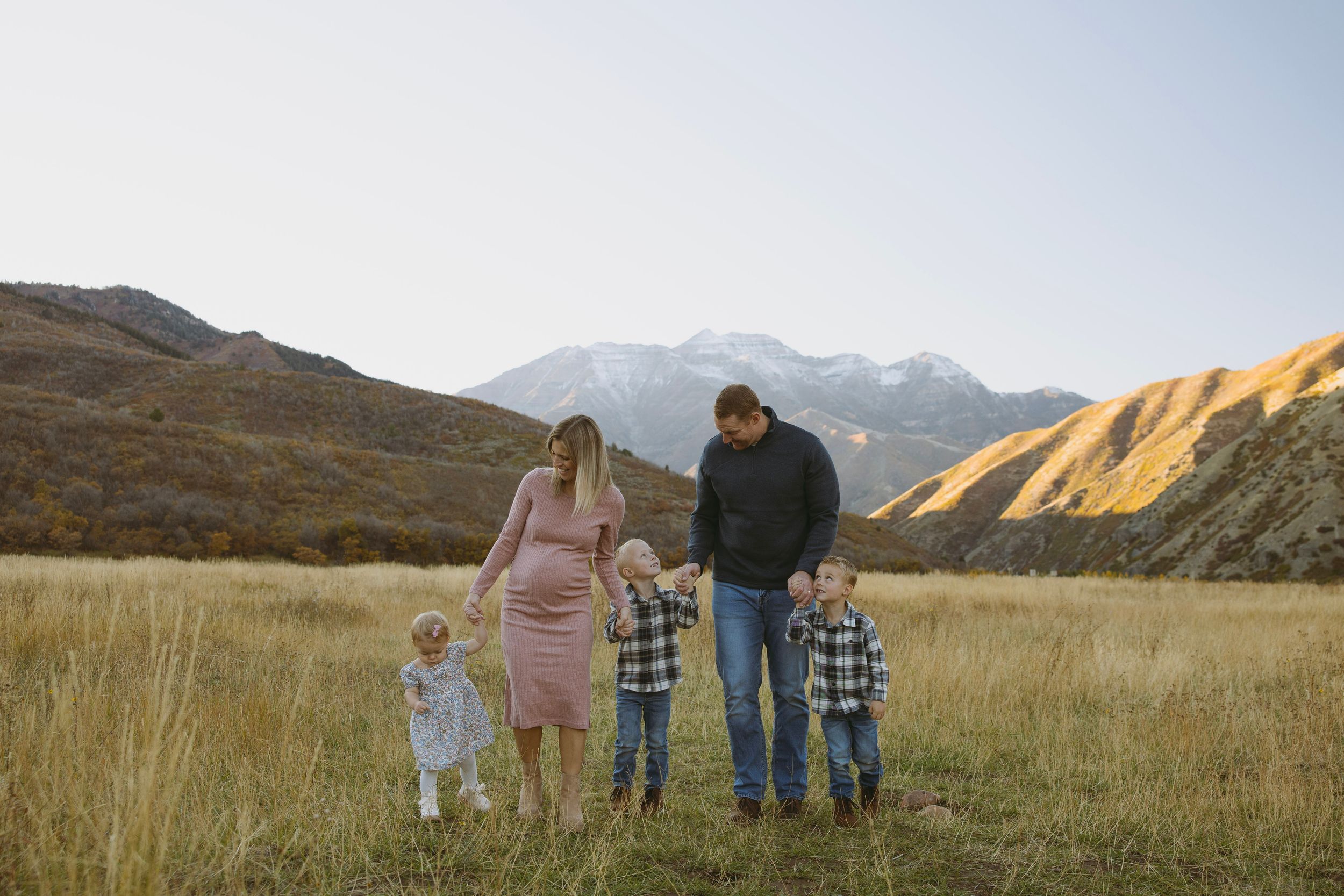 Family of five walking in a golden meadow with snow-capped mountains in the background during autumn.