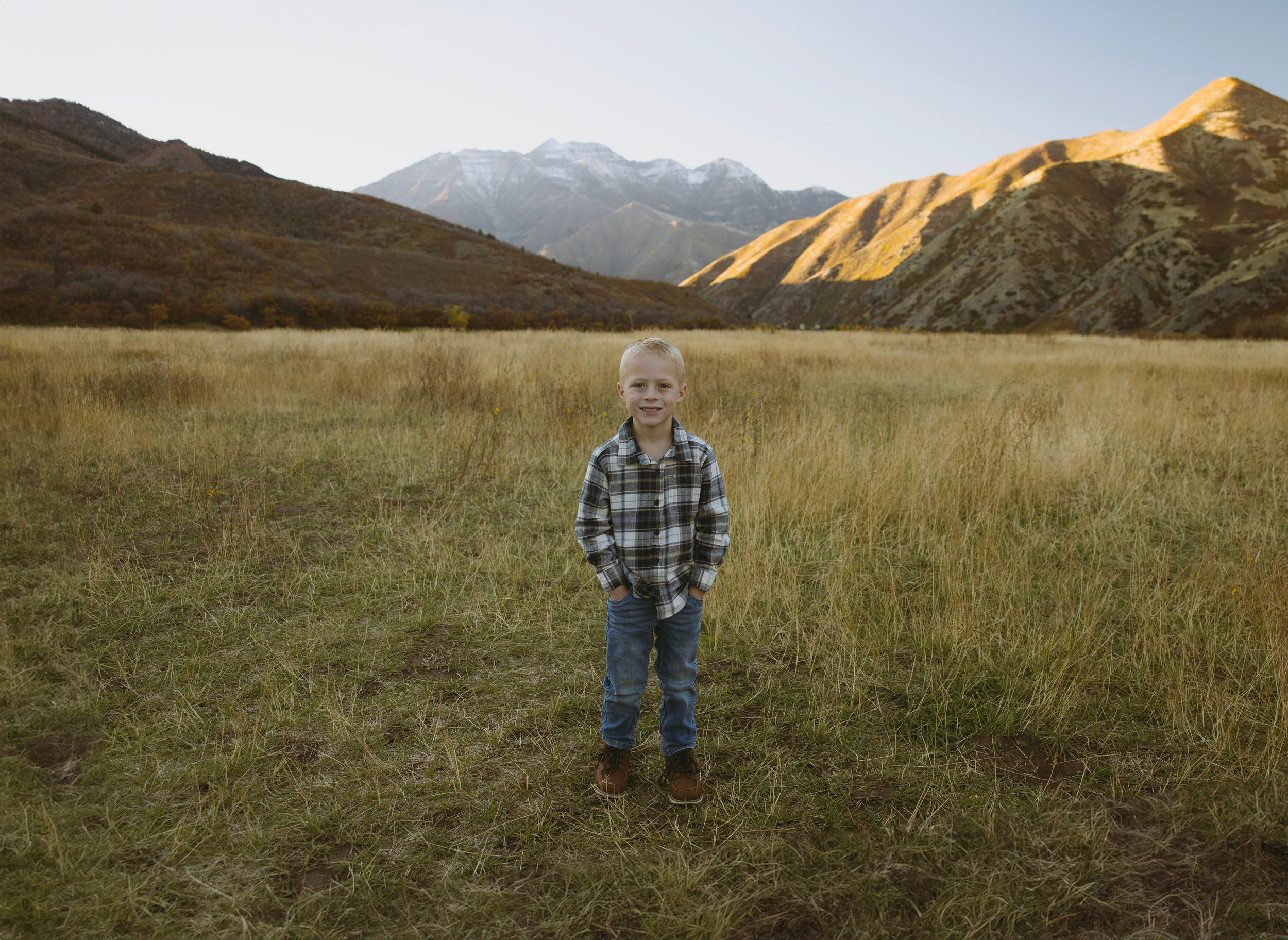 Young boy in plaid shirt standing in golden meadow with snow-capped mountains at sunset.