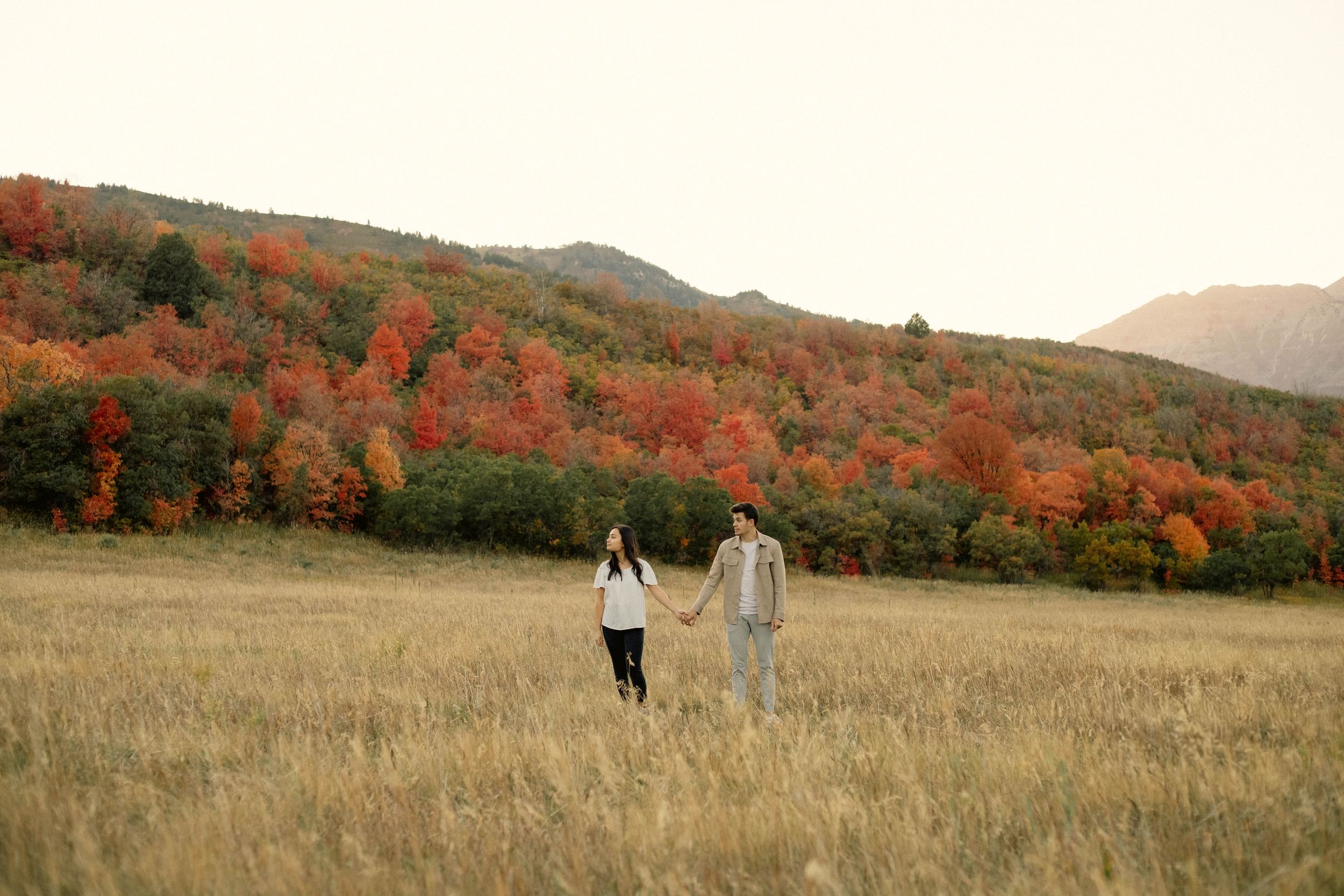 Couple holding hands walking through golden field with vibrant red and orange autumn foliage on hillside behind them.