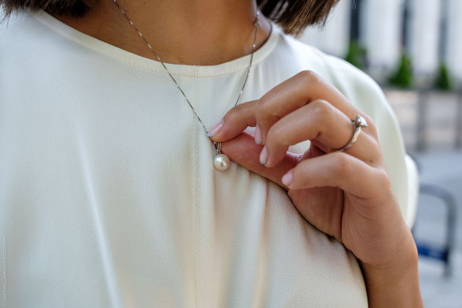 Woman in white top touching pearl pendant necklace, wearing a silver ring, close-up jewelry detail shot.
