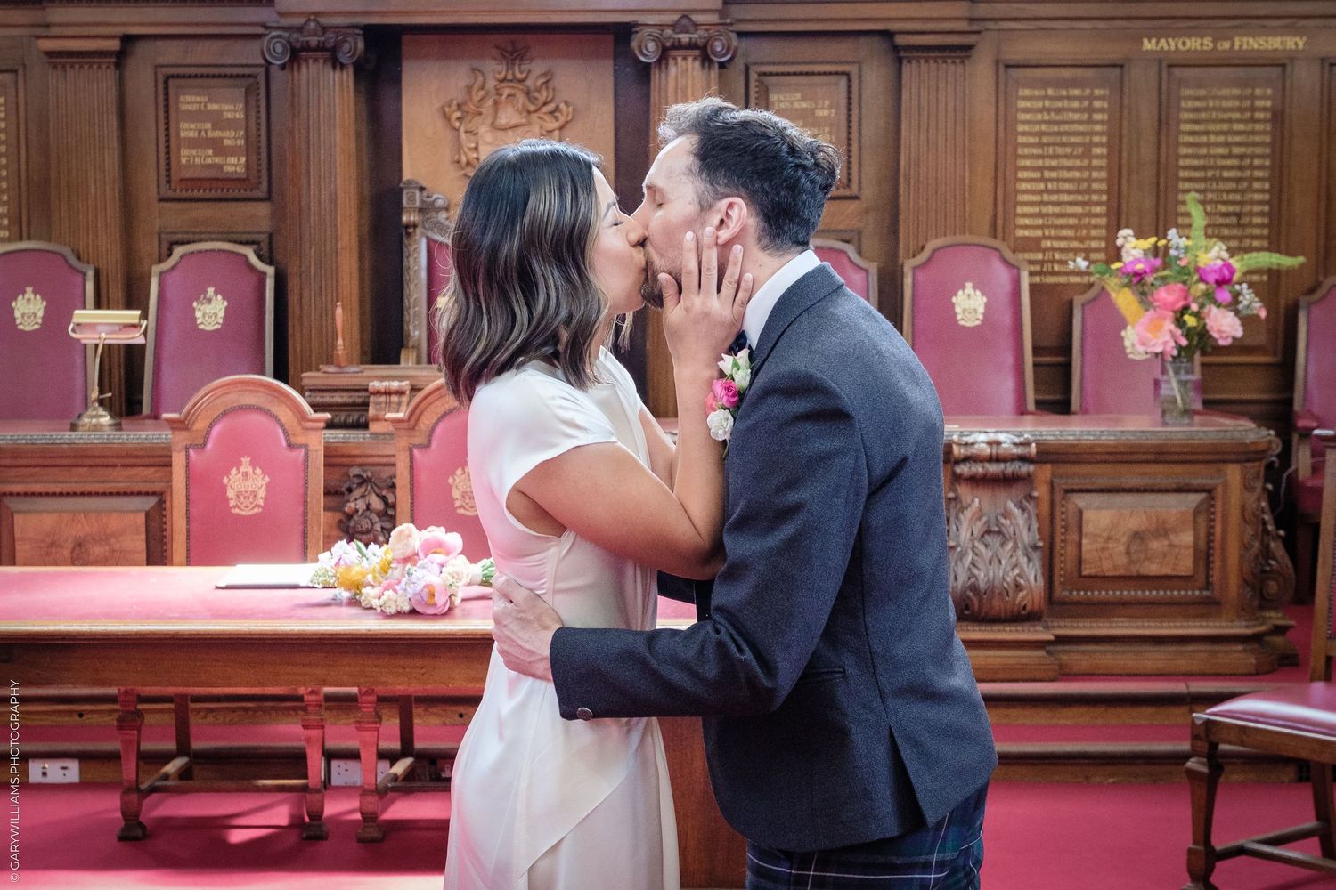 Couple sharing a first kiss at a civil wedding ceremony in an elegant wood-paneled registry office with pink flowers.