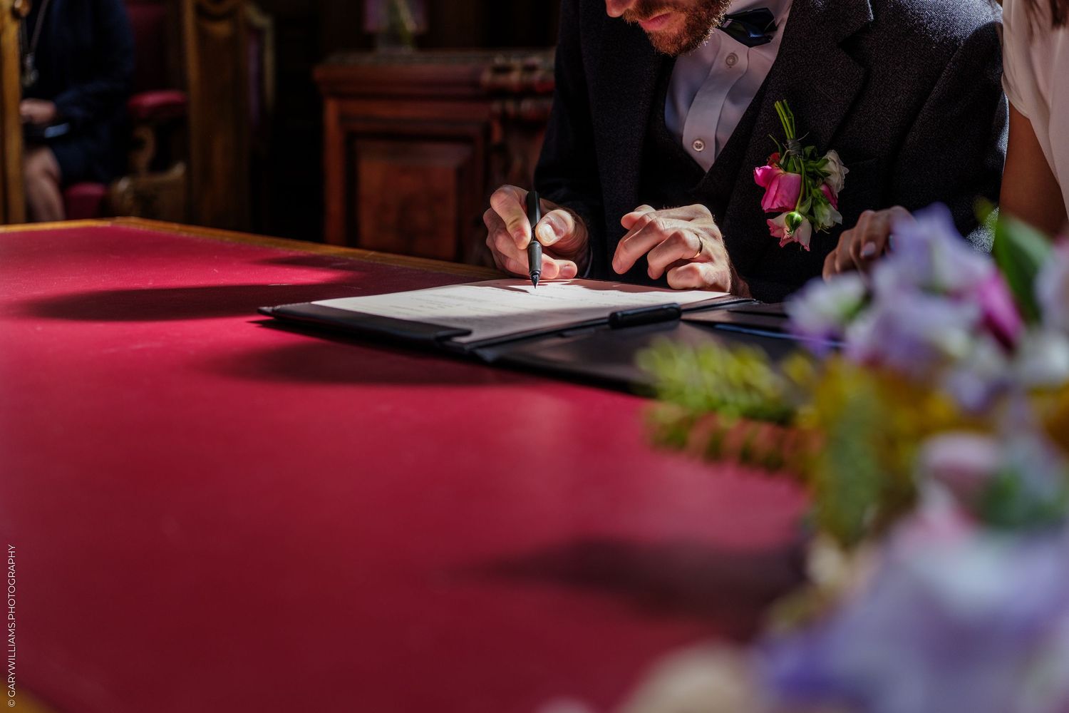 Groom signing wedding documents at a red velvet table adorned with flowers during a formal ceremony.
