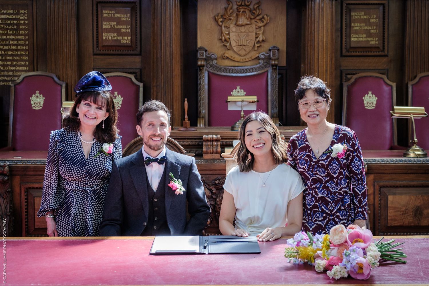 Couple signs wedding register with two witnesses in an ornate wood-paneled ceremony room with red decor.