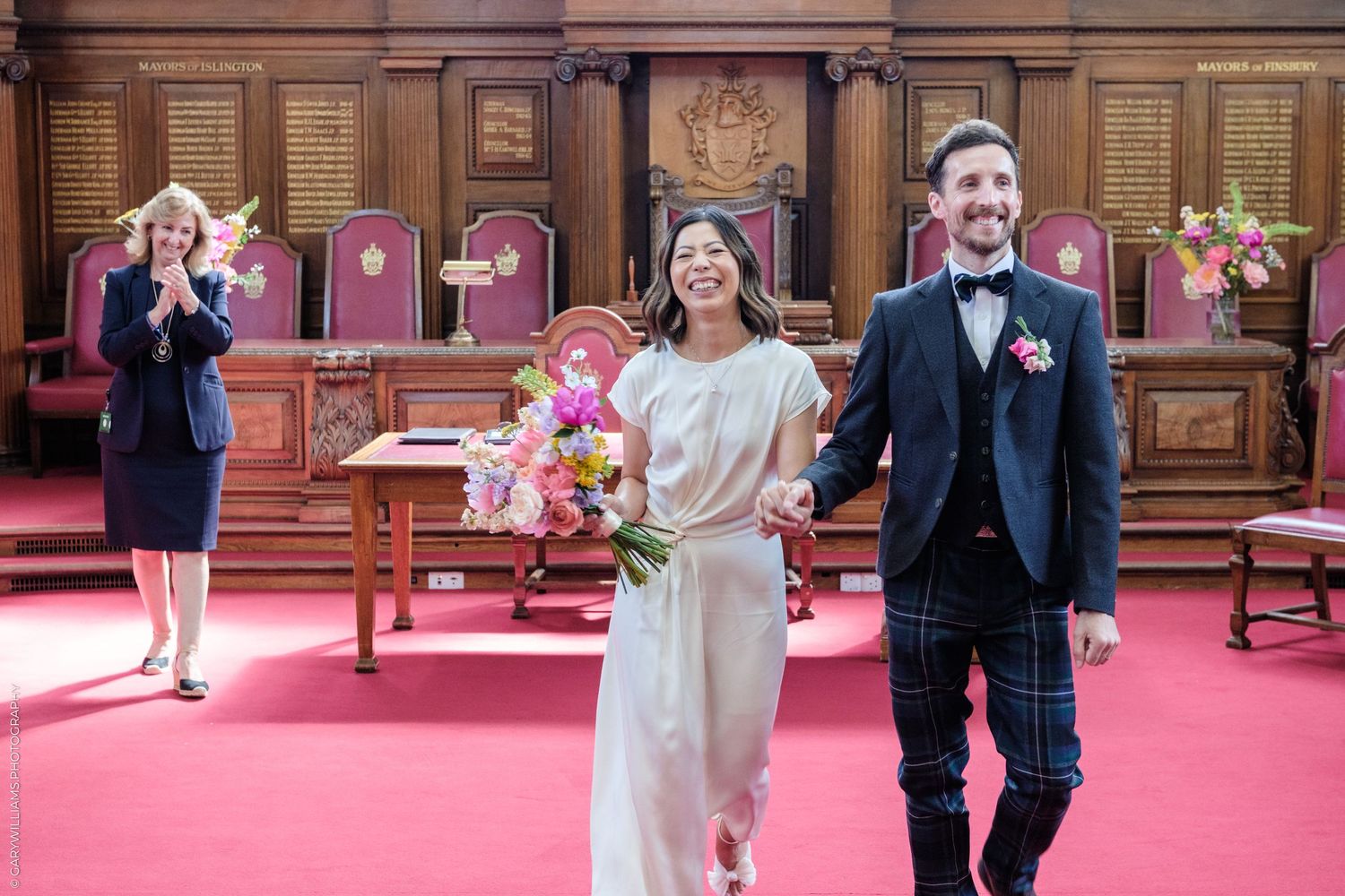 Joyful newlyweds walking hand in hand after civil ceremony in ornate red courtroom.