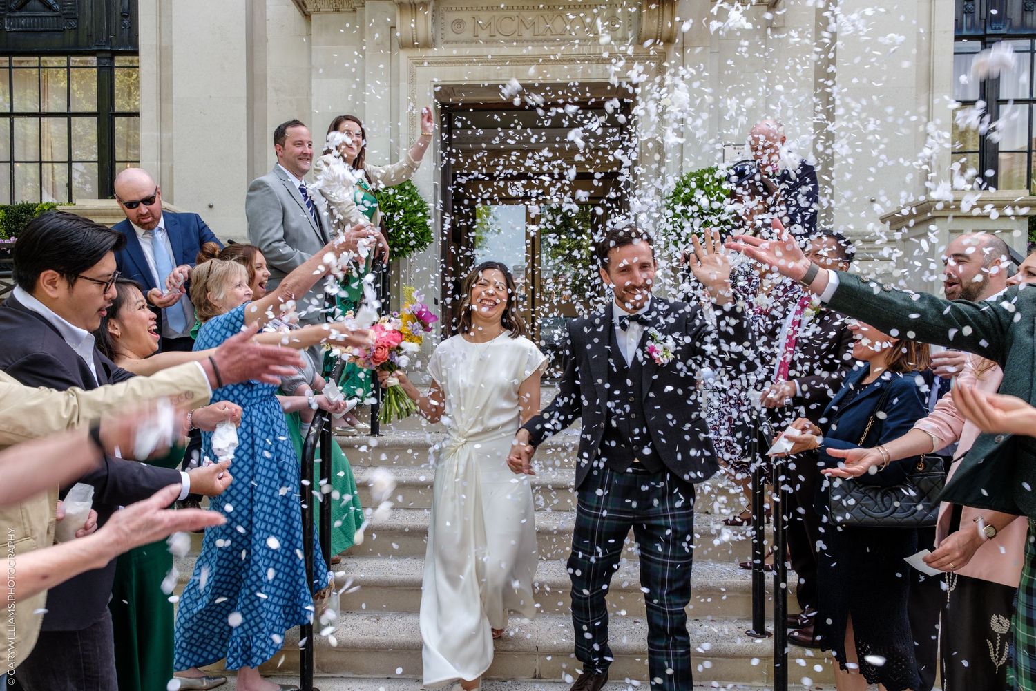 Joyful newlyweds exit wedding venue as guests shower them with white confetti outdoors.