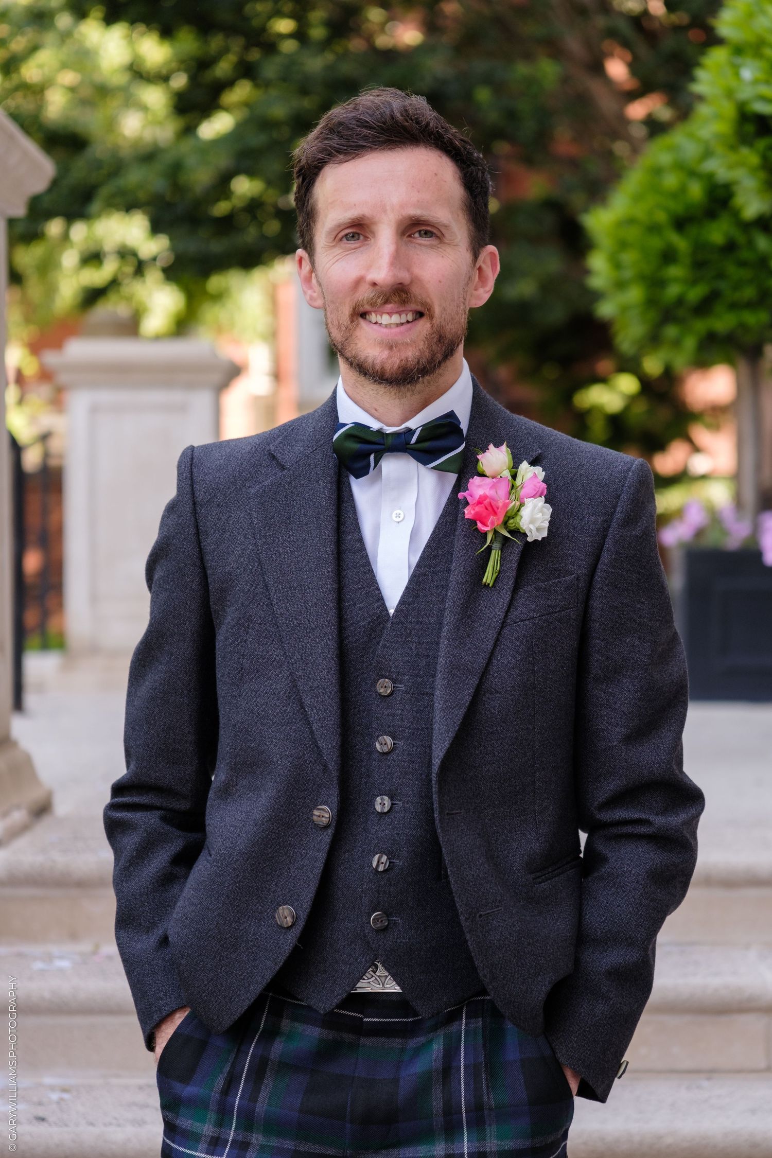 Smiling groom in tartan kilt, gray tweed jacket, waistcoat, bow tie, and pink floral boutonniere outdoors.