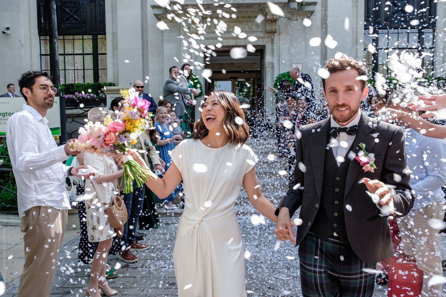 Joyful newlyweds walk through confetti shower outside venue, bride in white dress, groom in tartan kilt.