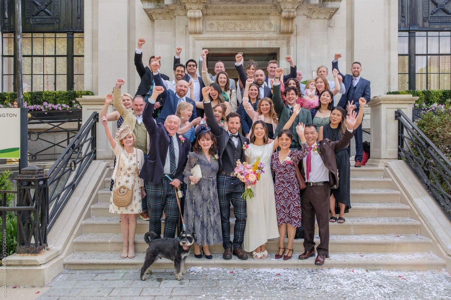 Wedding group photo on building steps, guests cheering with raised arms, bride holding bouquet, dog in foreground.