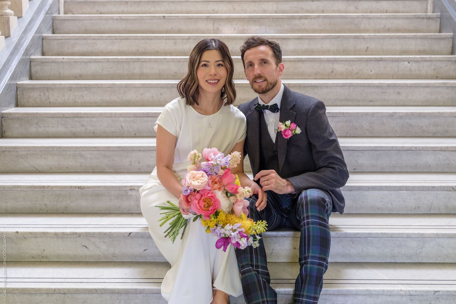 Bride and groom sitting on marble stairs, bride holding colorful bouquet, groom wearing plaid pants and bow tie.