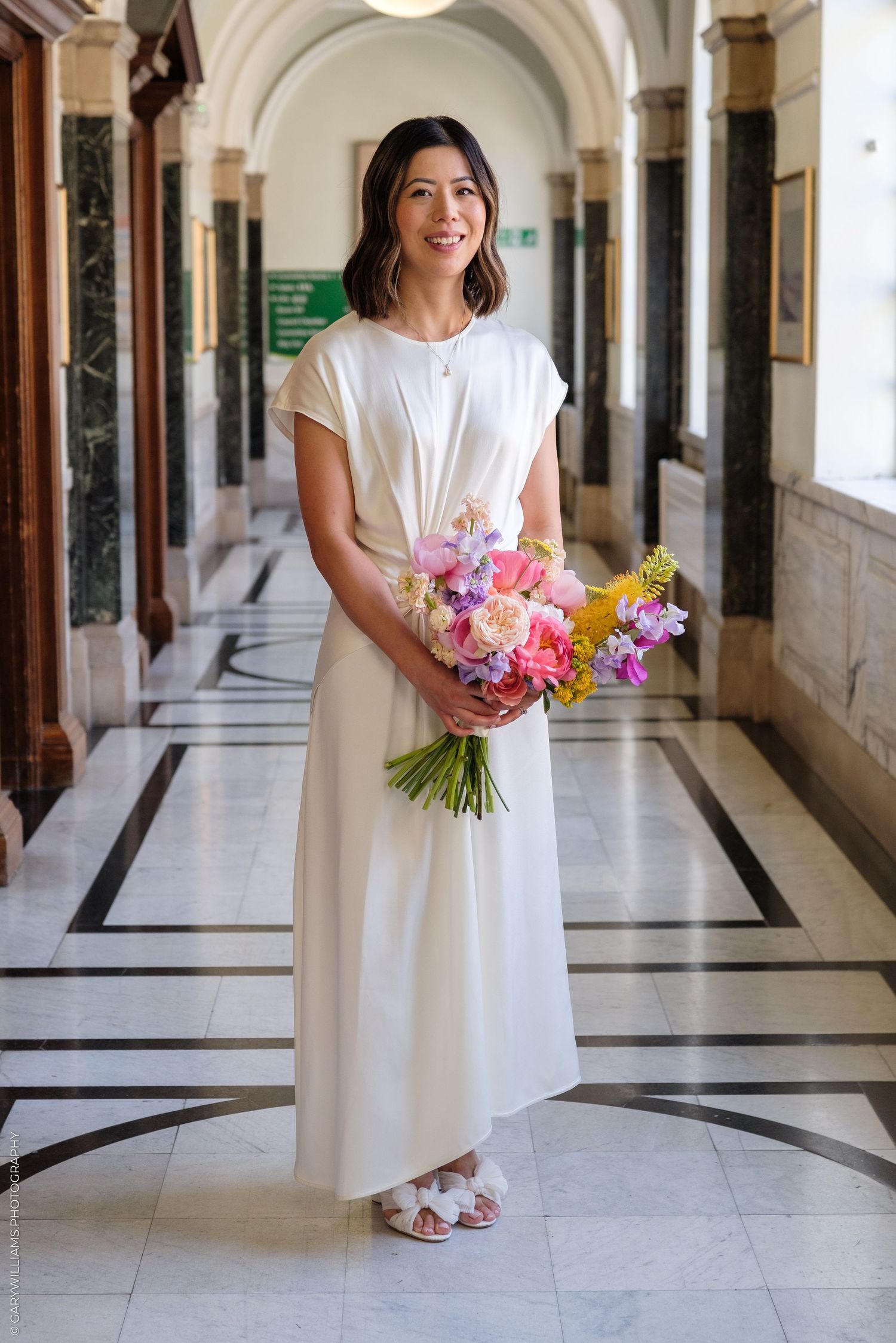Bride in white maxi dress holding colorful bouquet of pink, purple, and yellow flowers in an elegant marble hallway.