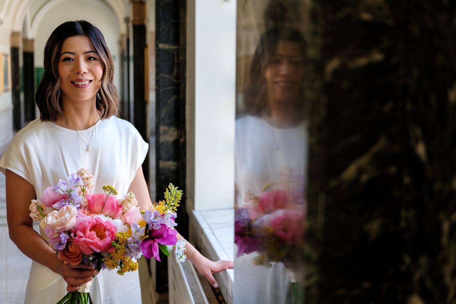 Woman in white dress holding colorful bouquet of pink, purple, and yellow flowers, standing in elegant hallway.
