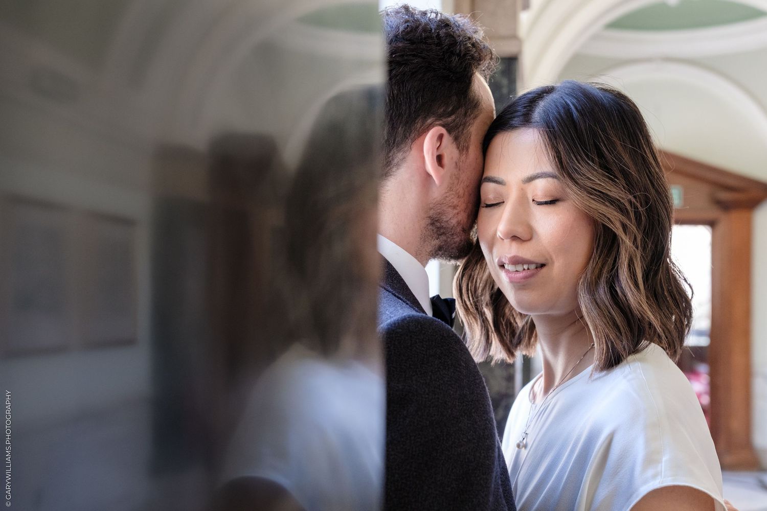 A couple shares an intimate moment, the woman smiling softly as the man leans close, reflected in a mirror.