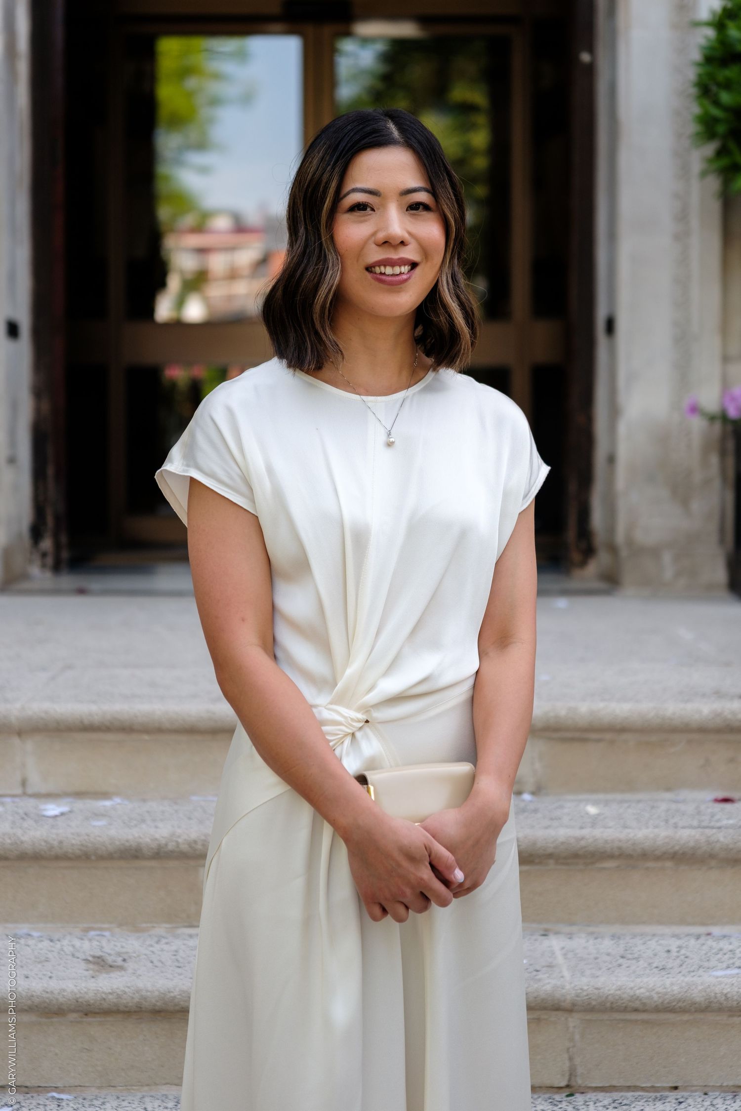 Smiling Asian woman in elegant white outfit standing on steps of a building.