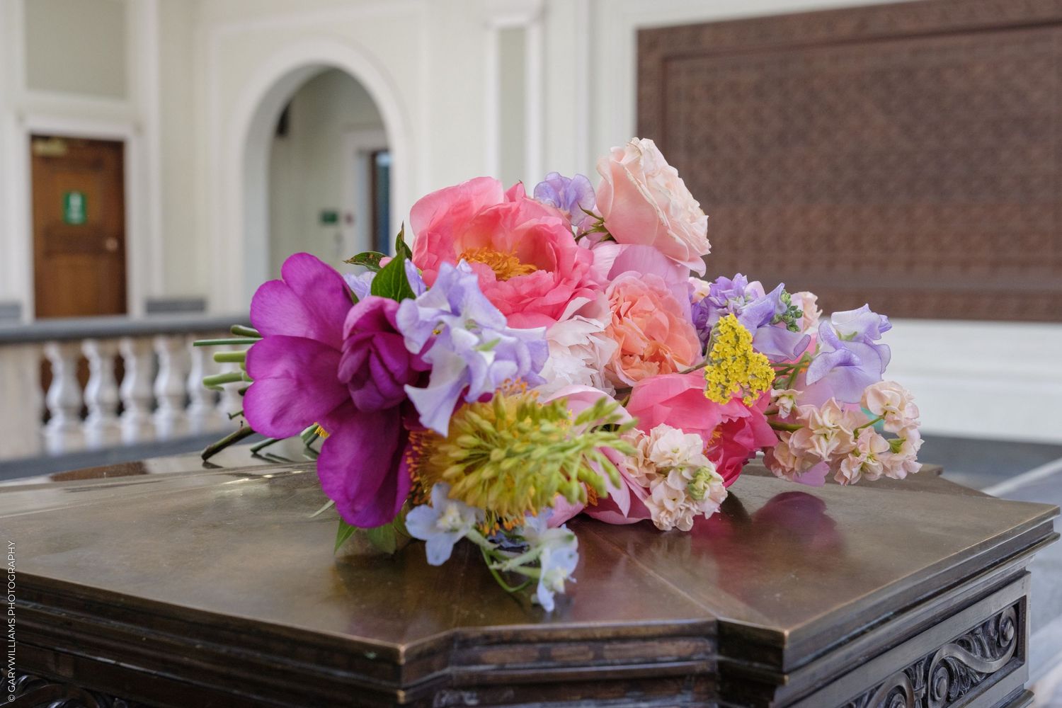 Colorful floral bouquet with pink peonies, purple flowers, and yellow blooms arranged on a wooden surface indoors.