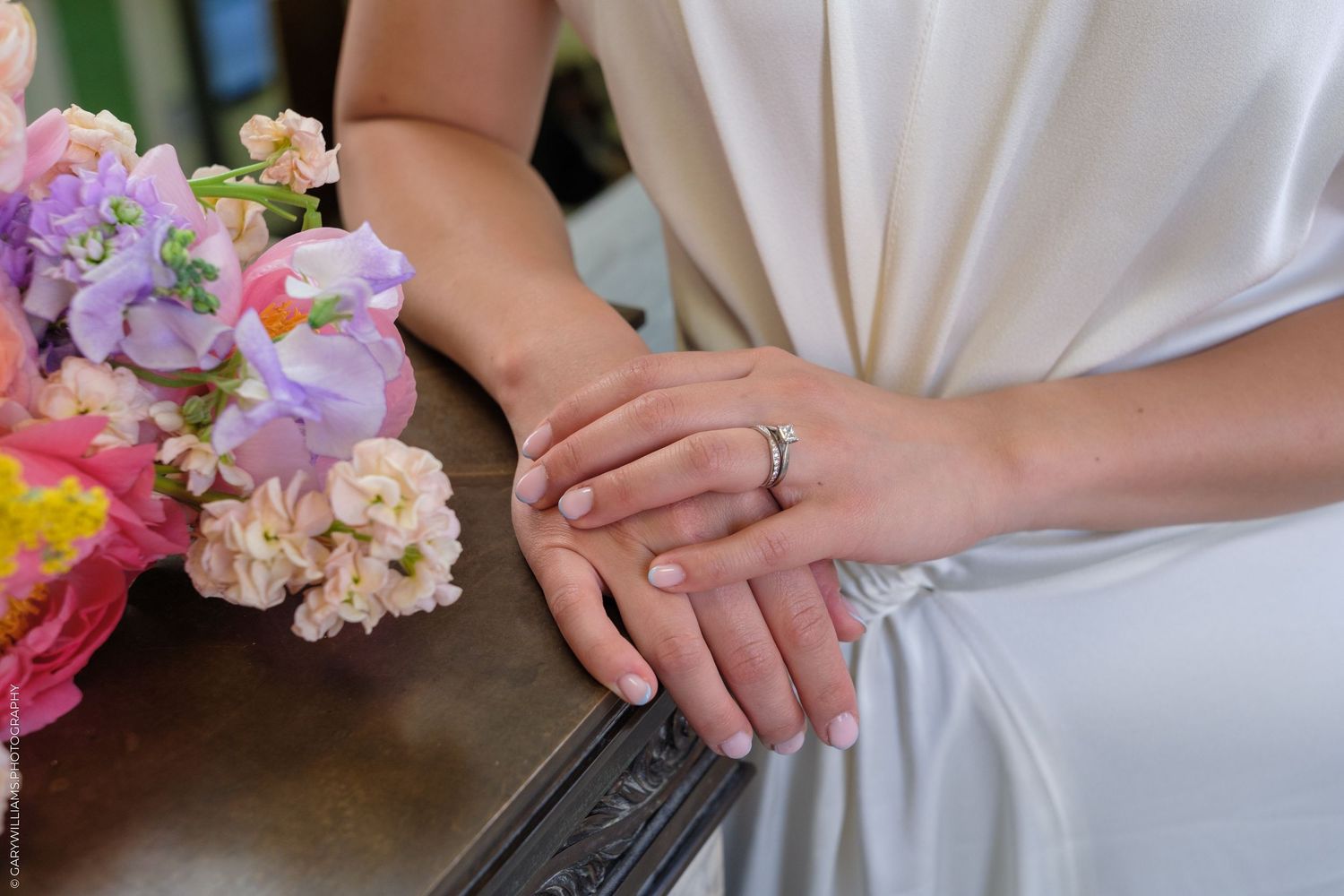 Close-up of bride's hands wearing a diamond ring resting on a dark surface beside a colorful floral bouquet.