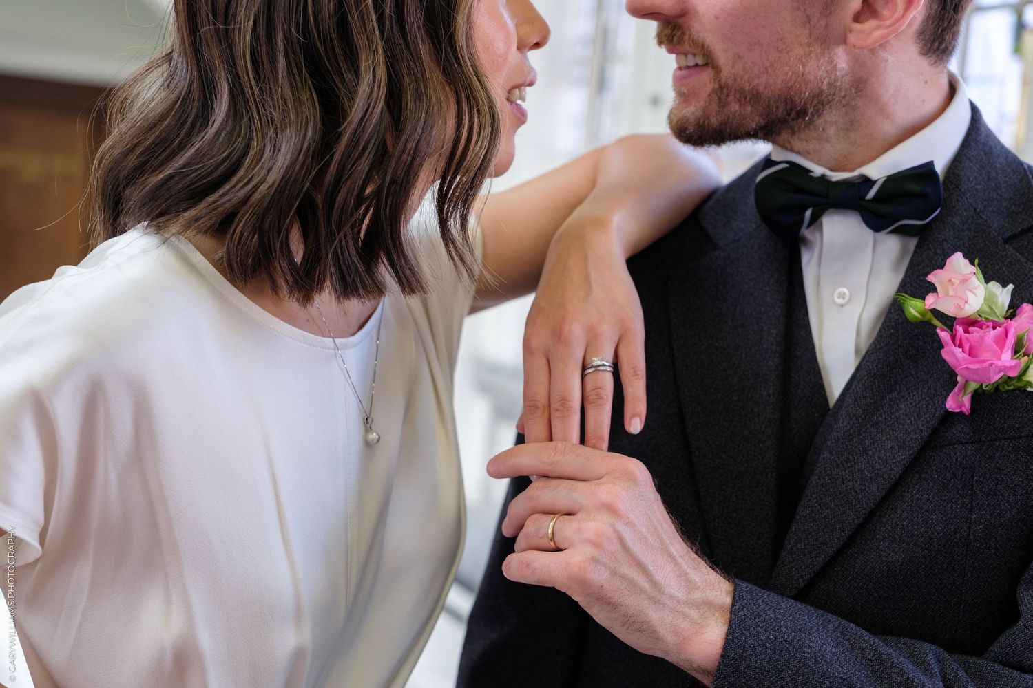 Newlywed couple exchanging smiles, bride wearing engagement ring in white dress, groom in dark suit with pink boutonniere.