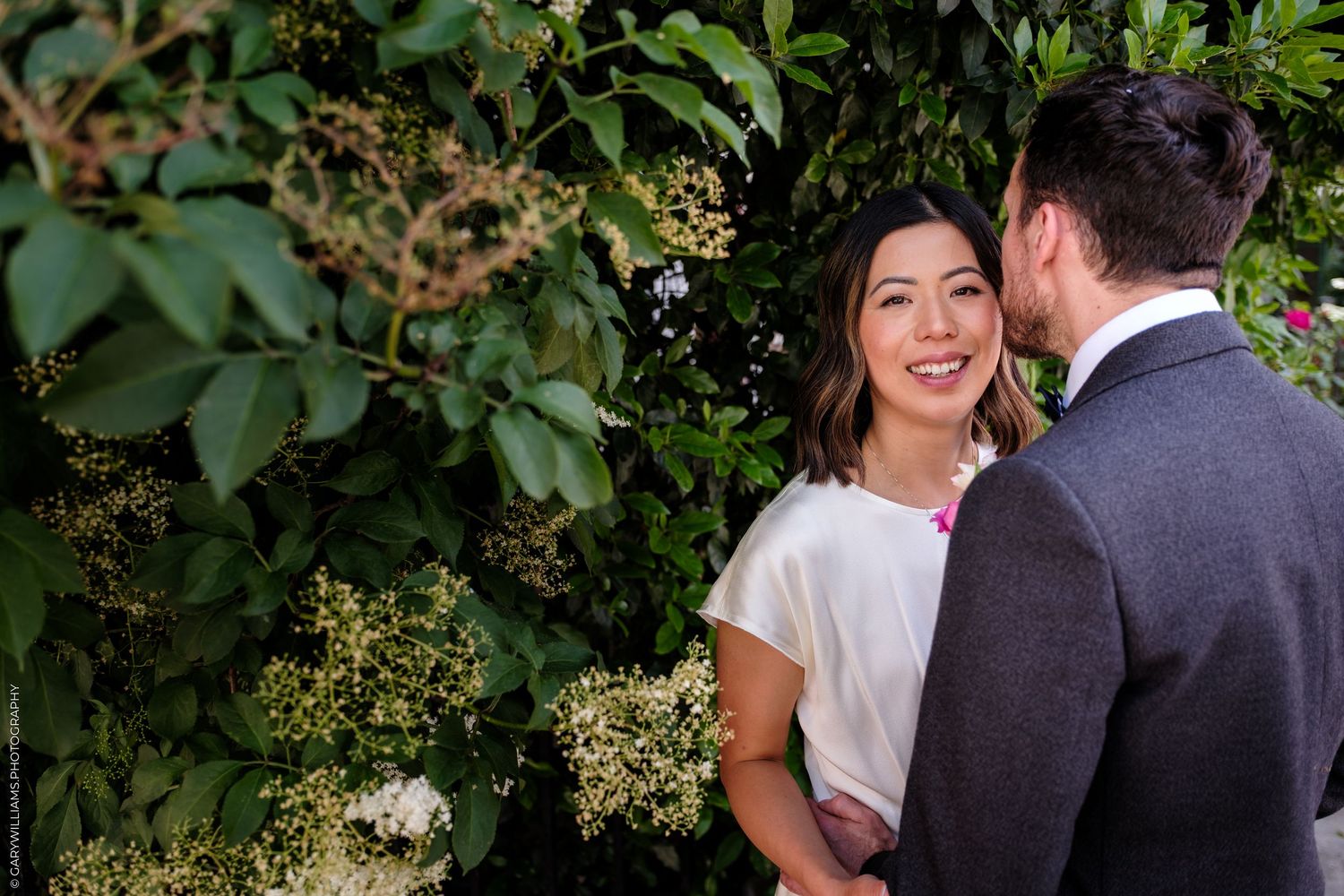 A smiling bride in a white dress stands with her groom among lush green foliage during an outdoor wedding.