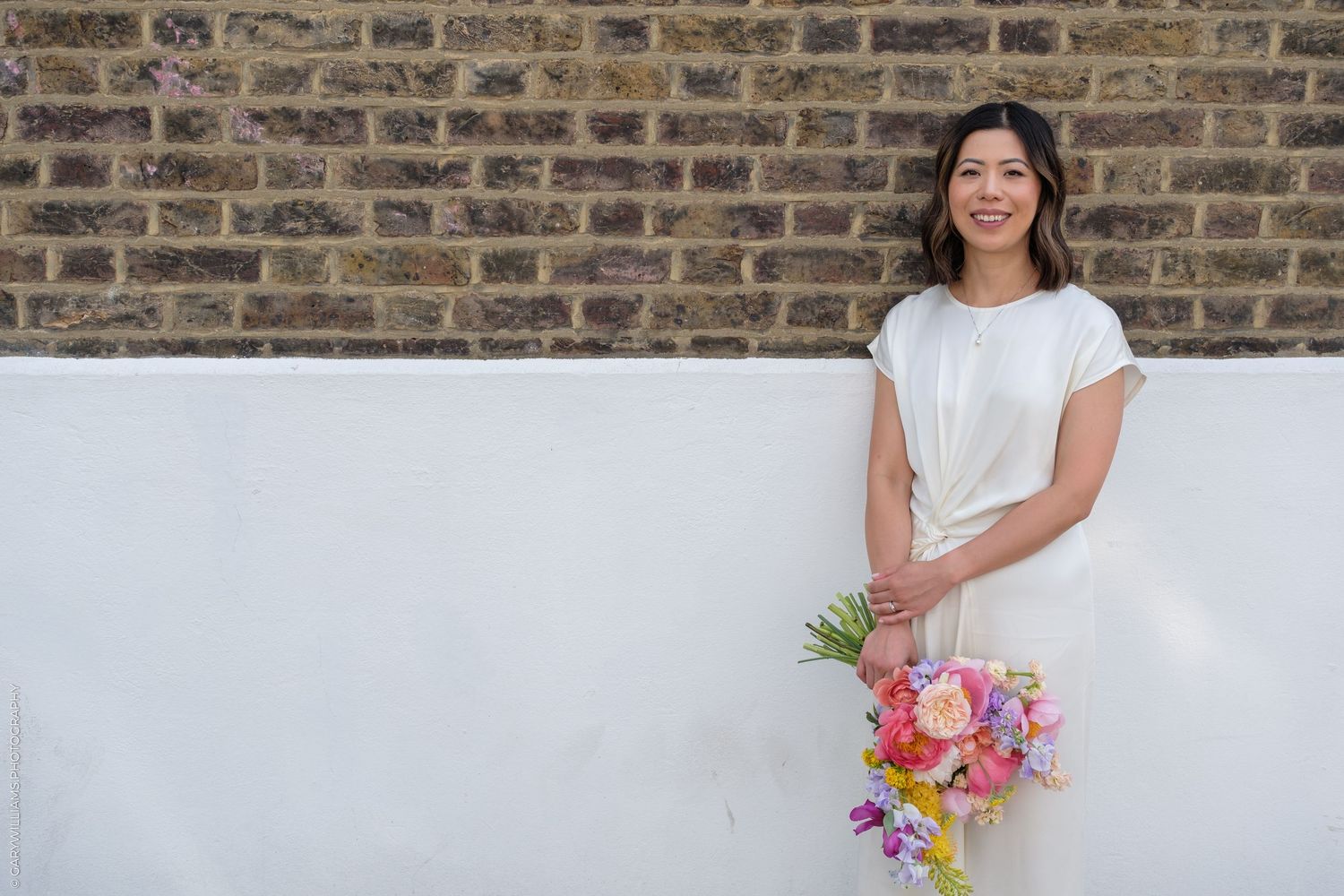 Smiling Asian bride in white dress holding colorful bouquet against brick wall.