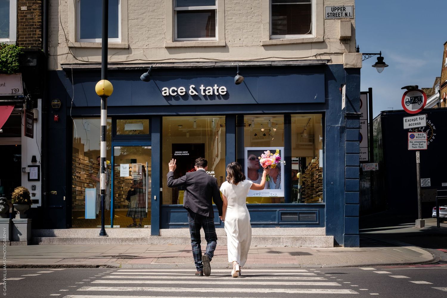 Couple crossing zebra crossing towards Ace & Tate opticians store on sunny day in London street.