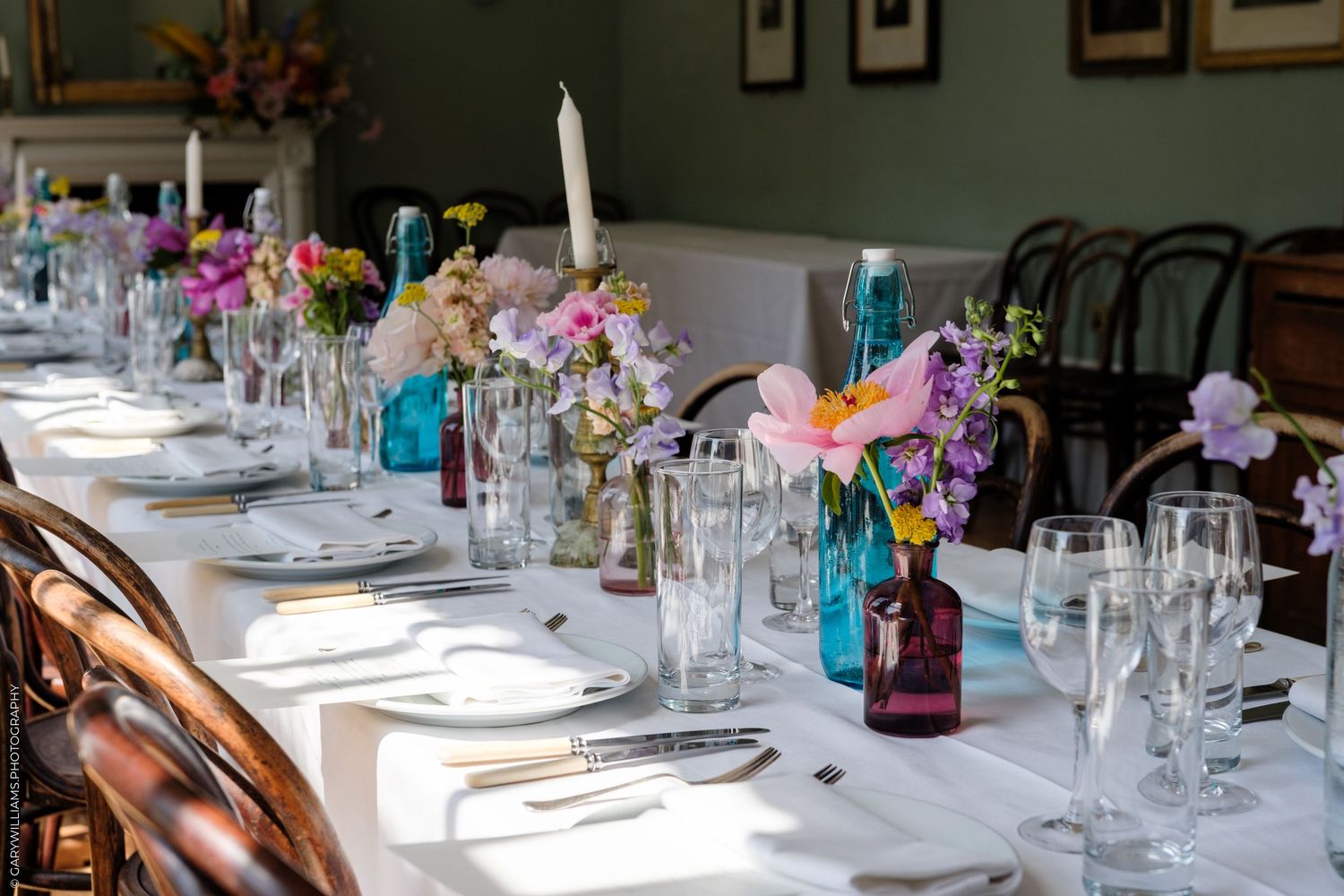 Elegant wedding reception table set with white linen, colorful flowers in blue and purple vases, candles, and glassware.