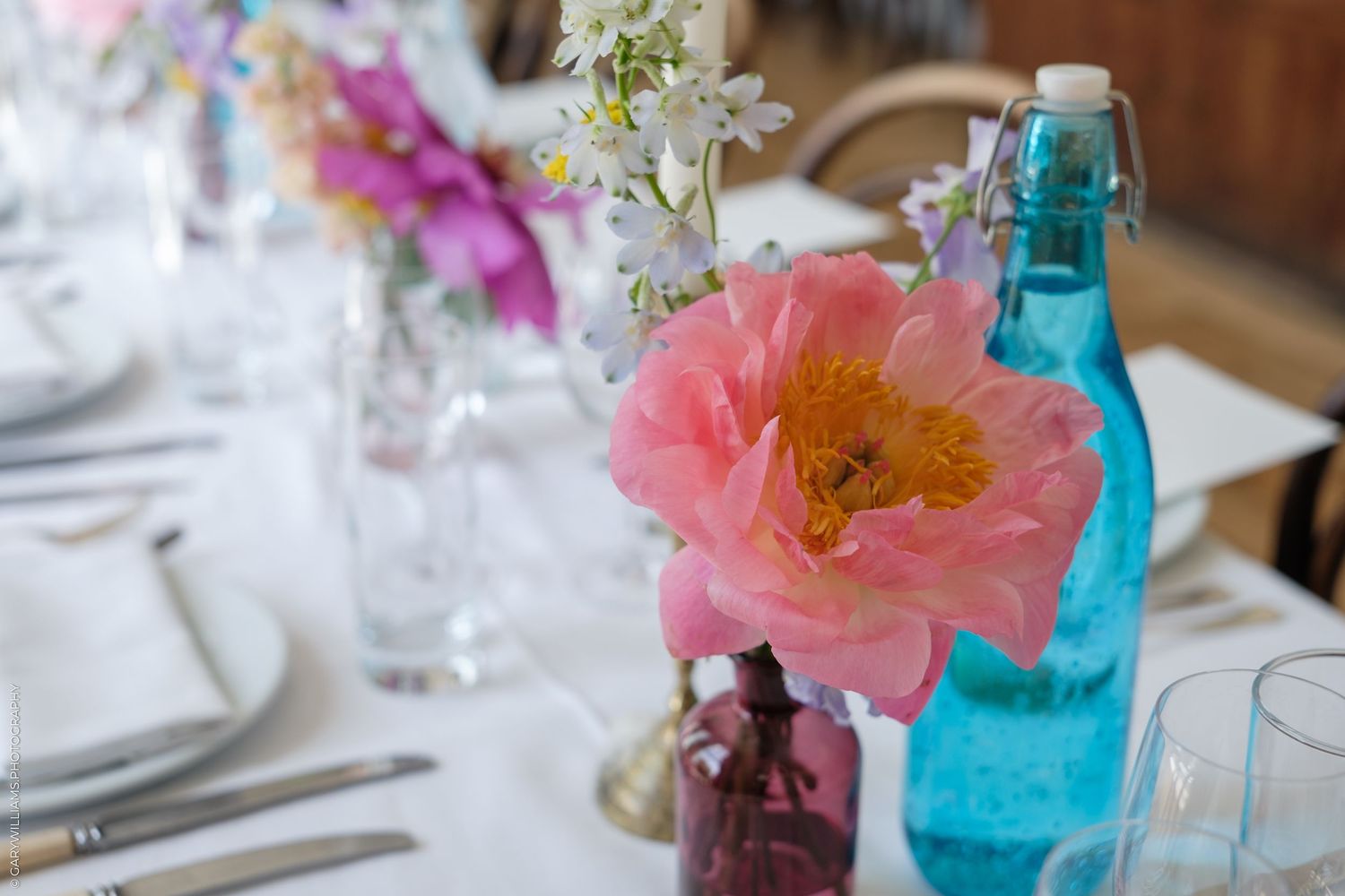 Elegant wedding table with pink peony, blue glass bottle, and delicate floral arrangements on white linen.