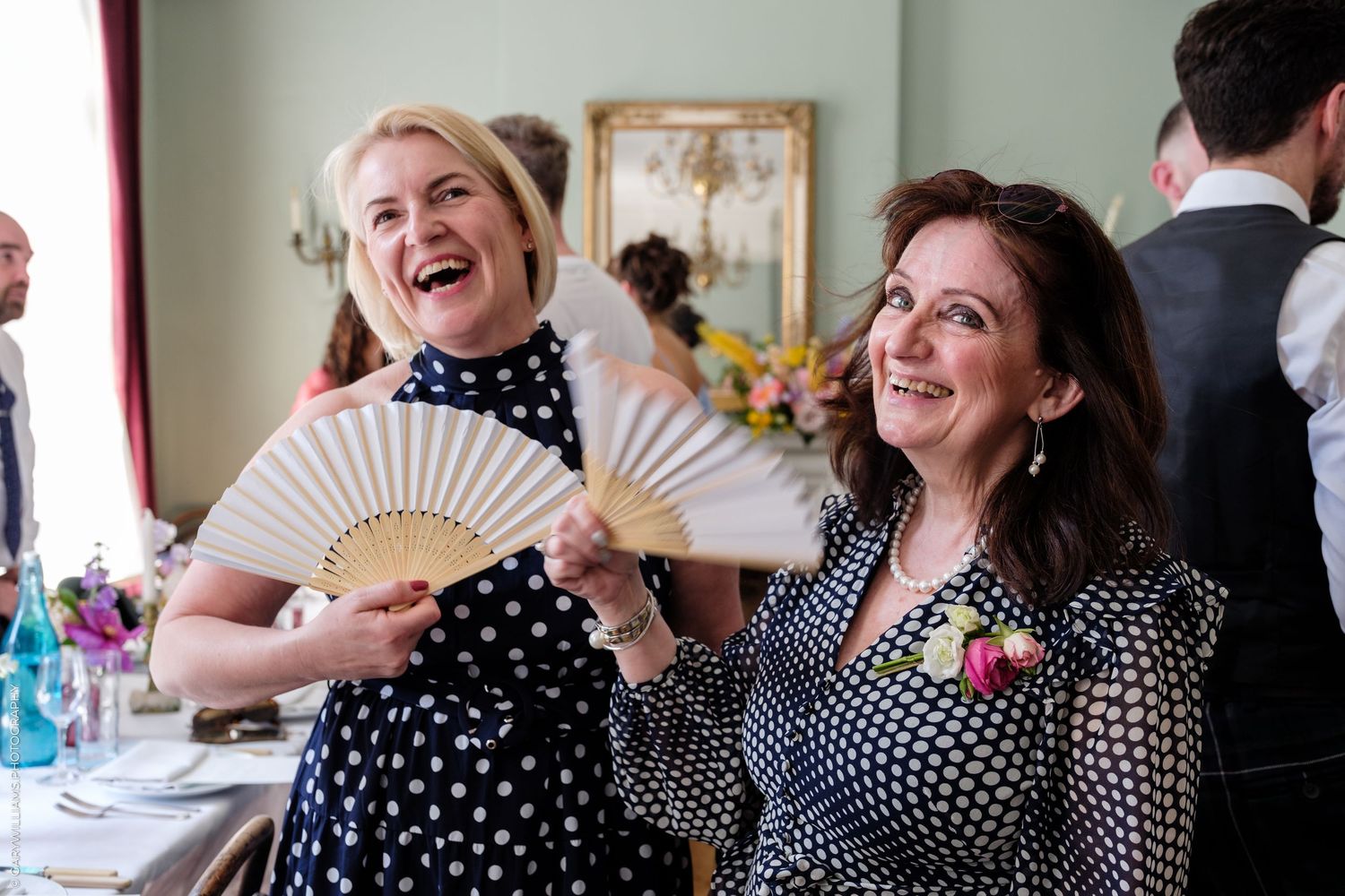 Two women laughing and holding decorative hand fans at a wedding reception in a green-walled venue.